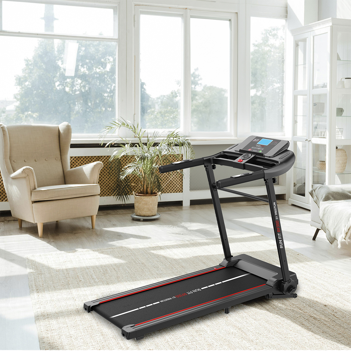 Czarna bieżnia w jasnym pomieszczeniu z dywanem, krzesłem i oknami. Roślina w tle.