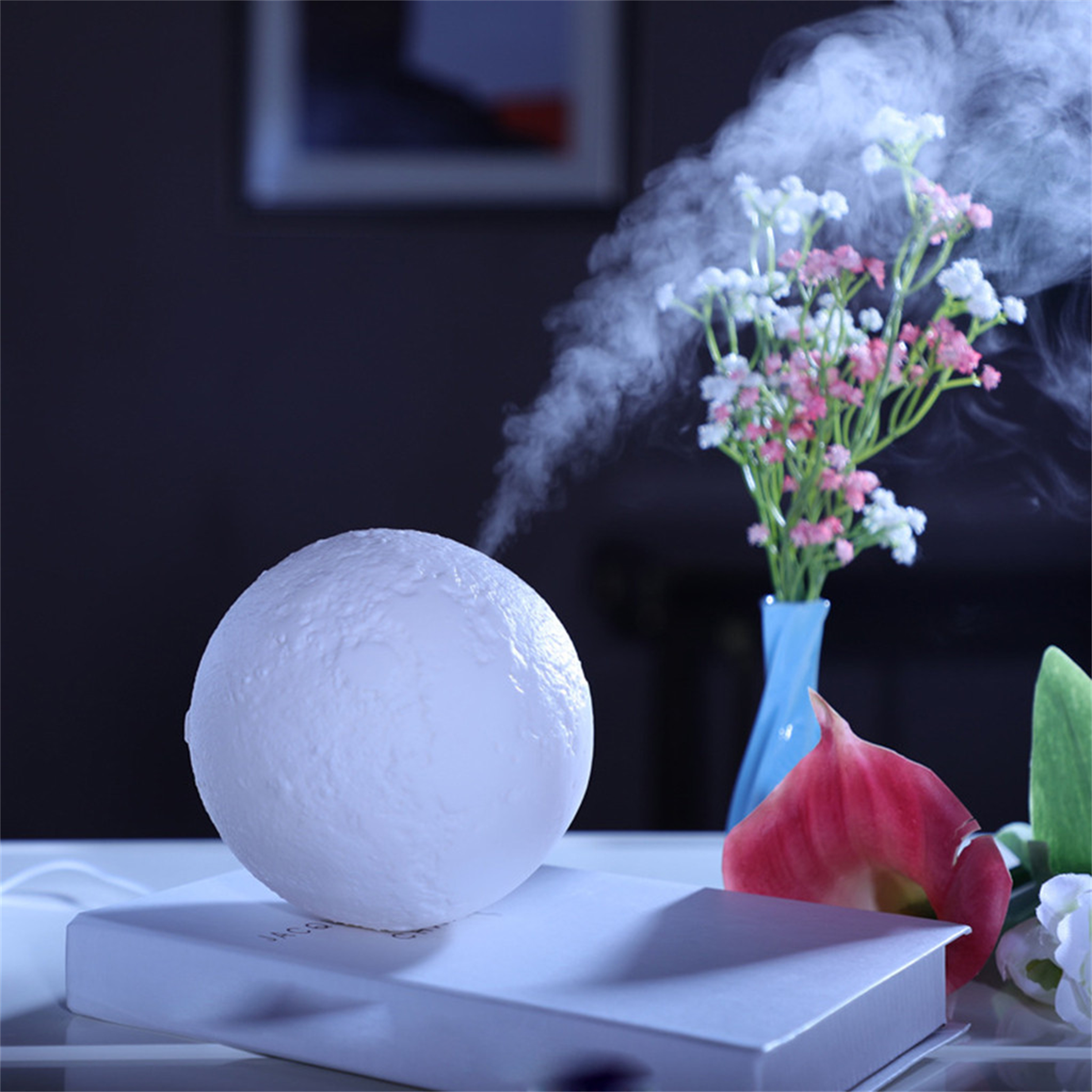 Mondförmige Lampe mit Dampf, der daraus austritt. Blumen in einer Vase und ein Buch sind in der Nähe.