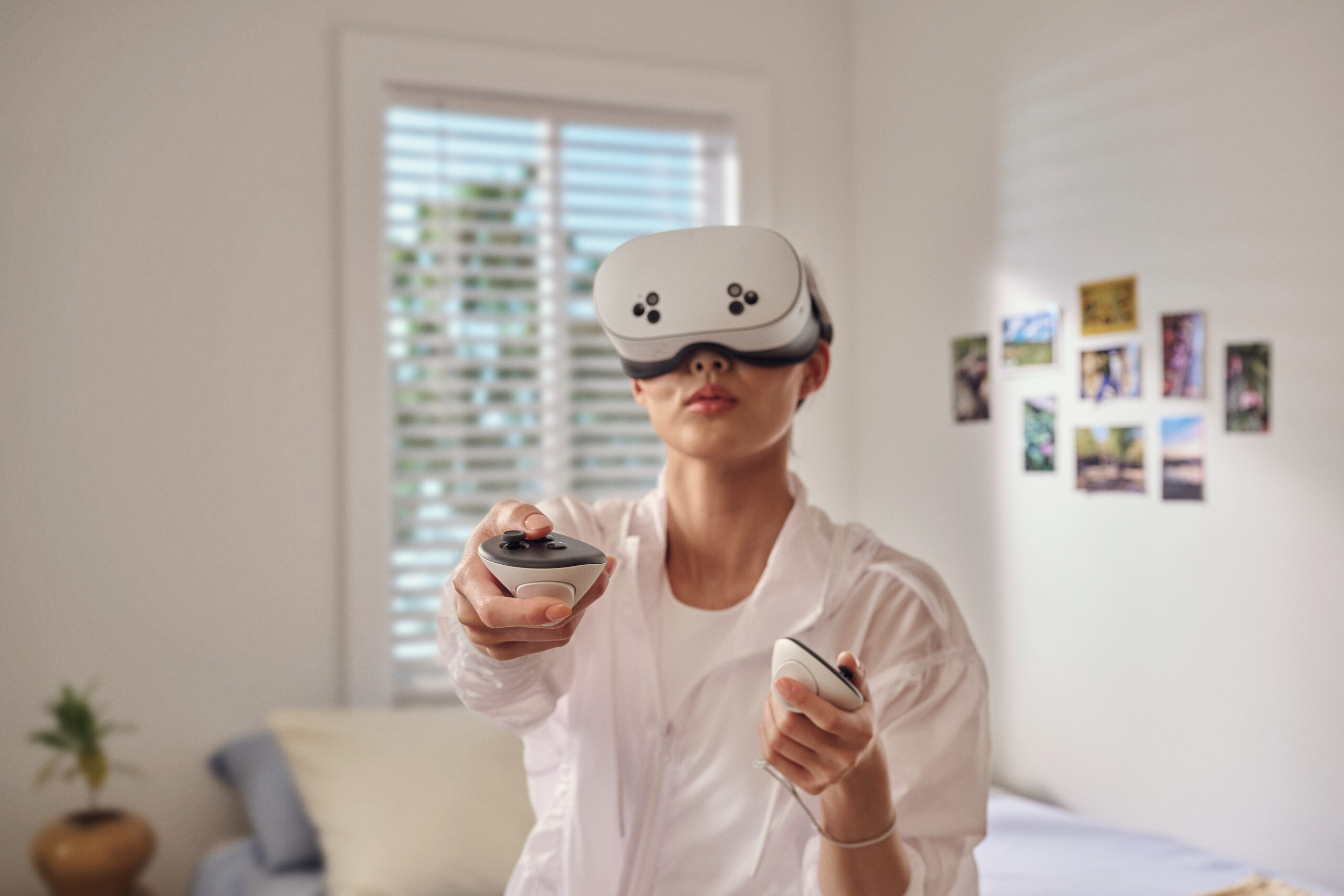 Una persona che indossa un visore VR, con controller. Sono in una stanza con una finestra e foto sul muro.