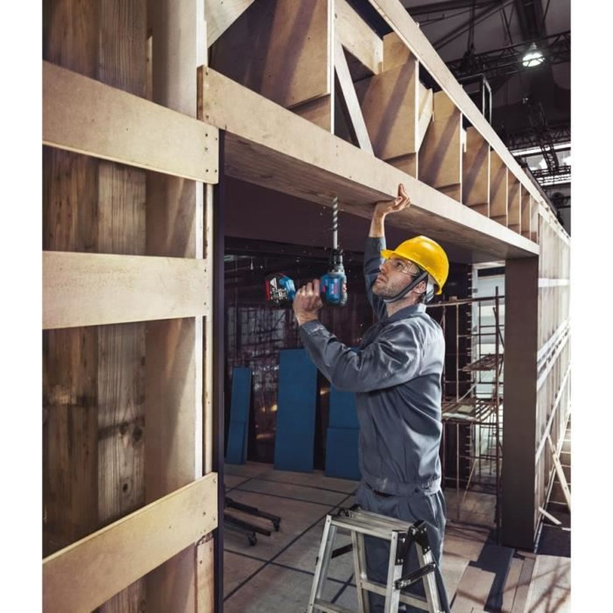 Un hombre con casco amarillo taladra madera. Está en una escalera de mano, en una estructura de madera.