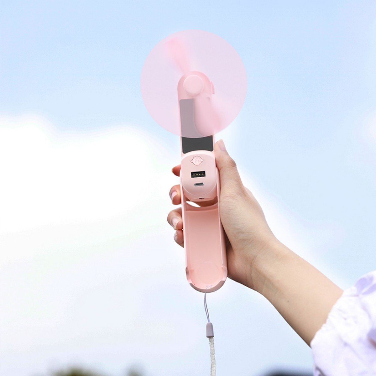 Die Hand einer Person, die einen rosa Mini-Ventilator vor einem blauen Himmel mit Wolken hält.