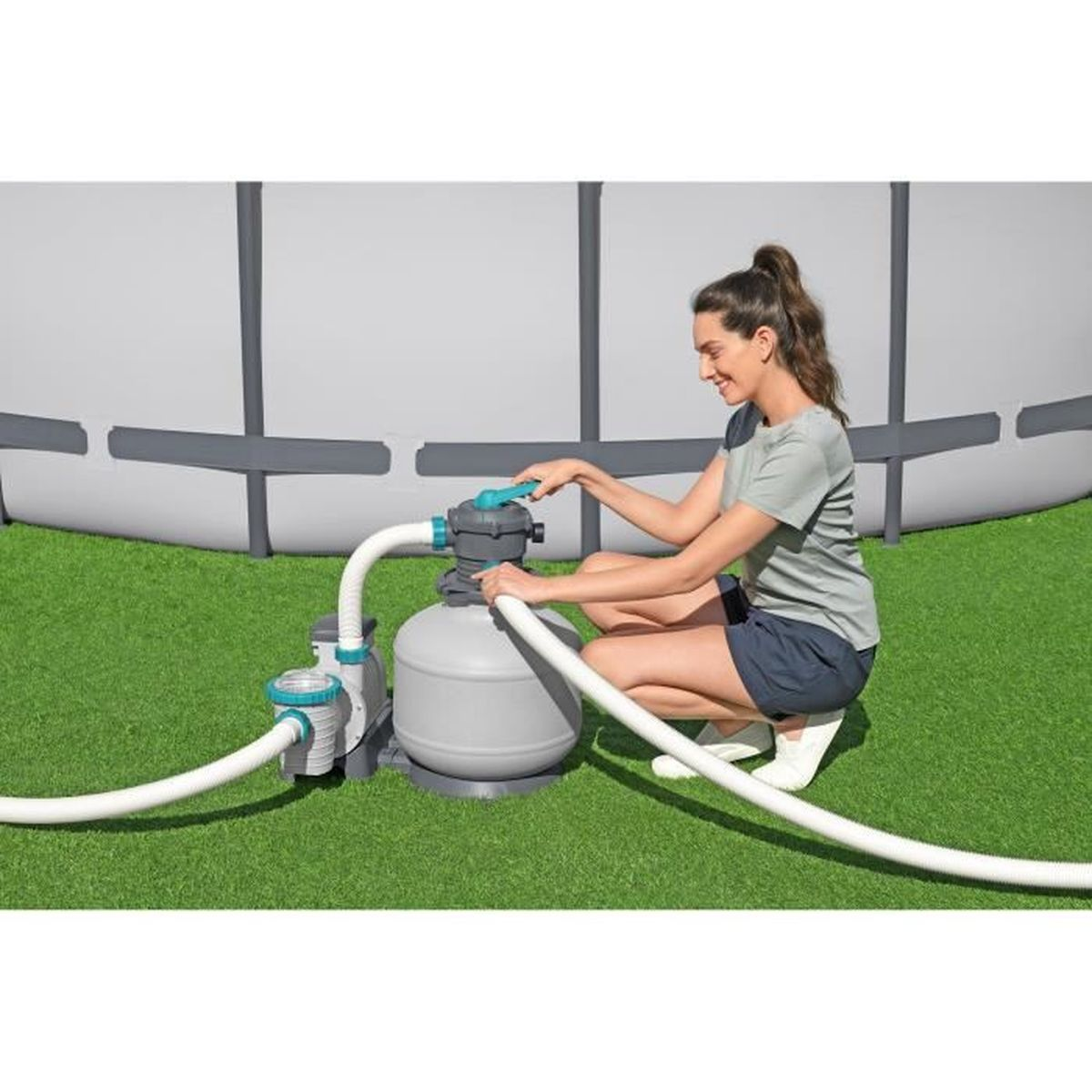 Kobieta klęczy przy filtrze basenowym, obsługując go na zielonej trawie.