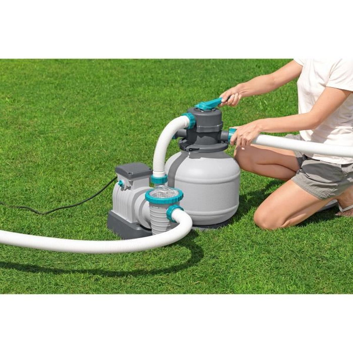 Osoba obsługująca filtr pompy basenowej na zielonym trawniku. Pompa basenowa jest szara i turkusowa.
