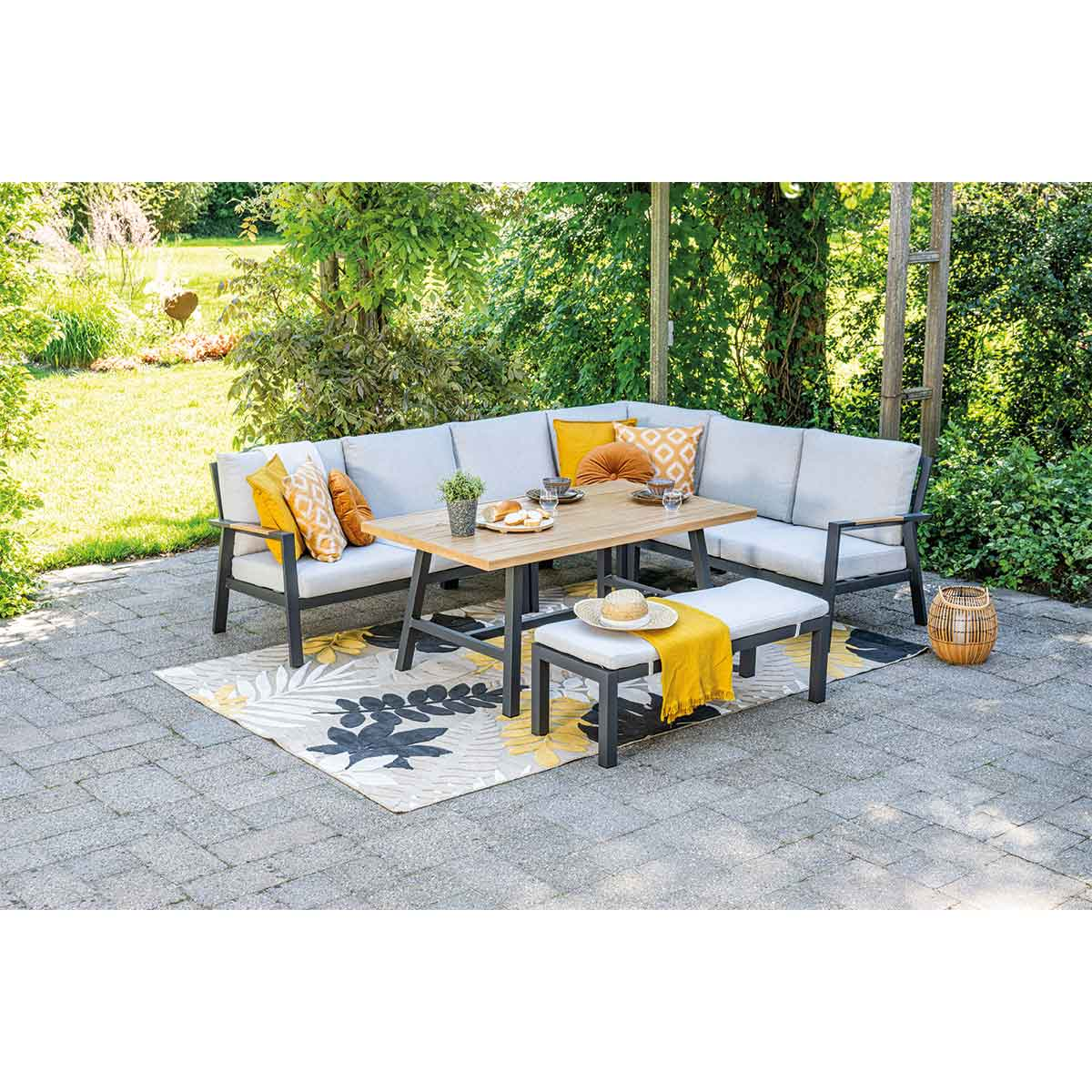 Buitenscène met hoekbank en tafel. Op tafel staan eten en drinken. Vloerkleed en decoratieve items.