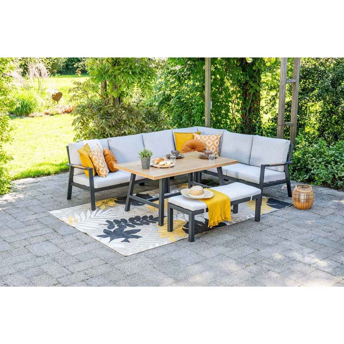 Salon de jardin avec table, banc et coussins sur un tapis à motifs, dans un jardin.