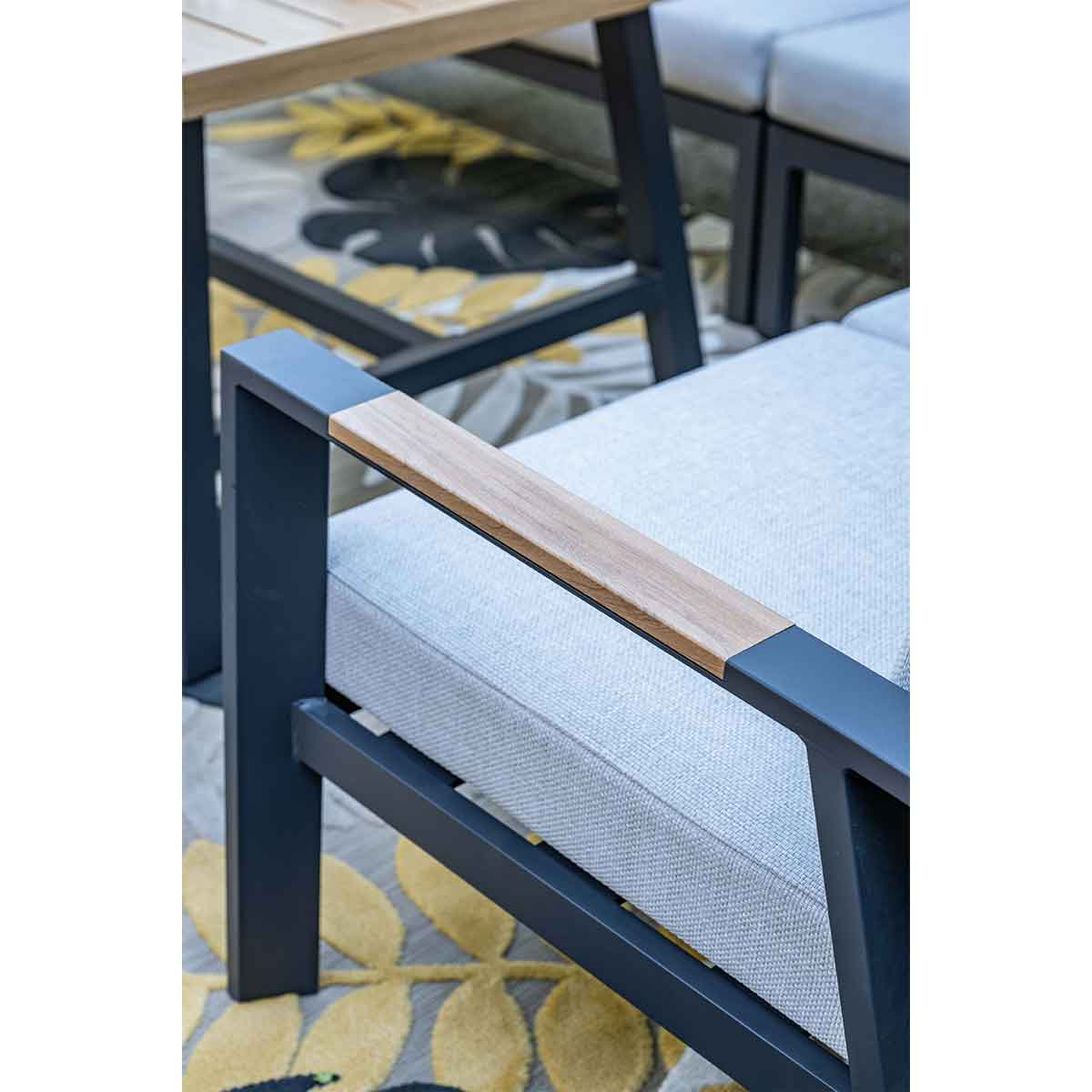 Gros plan sur un accoudoir en bois sur un canapé d'angle d'extérieur. Des coussins gris et un cadre en métal foncé sont visibles.
