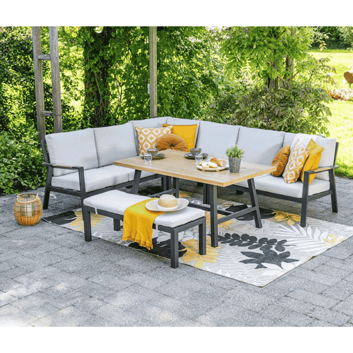Coin salon extérieur avec canapés gris, table en bois et banc. Coussins jaunes et oranges, tapis et plantes.