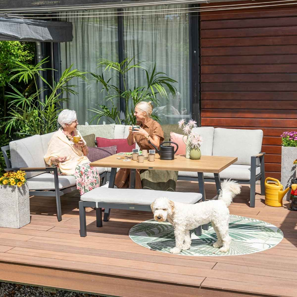 Deux femmes assises à une table extérieure avec un chien debout à proximité. Aménagement de mobilier extérieur.