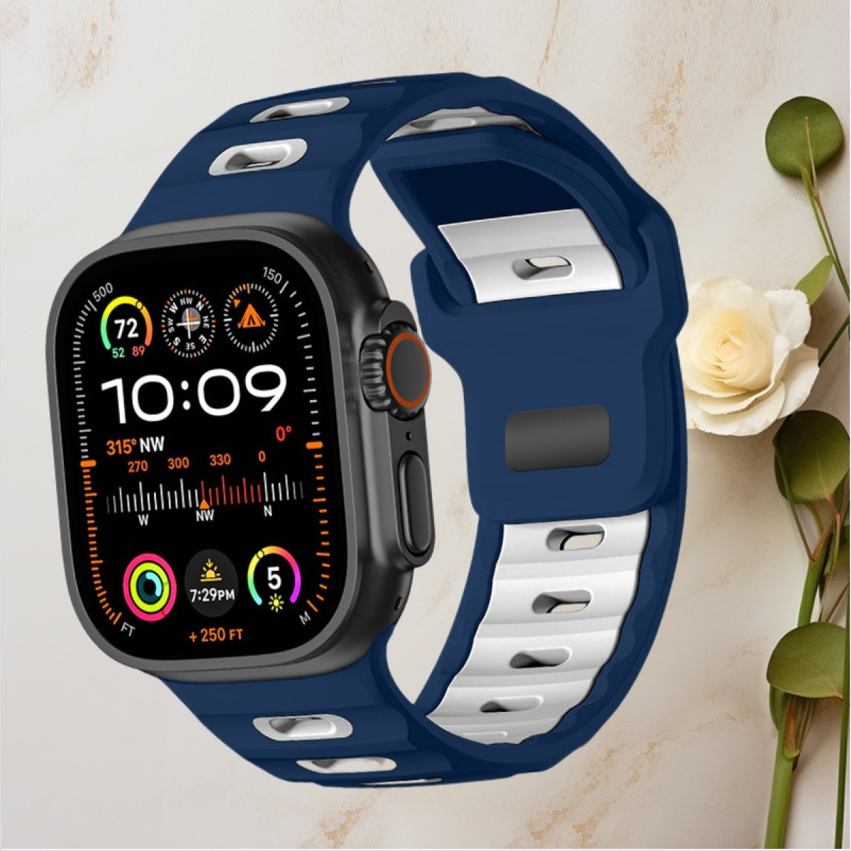 Smartwatch nero con cinturino blu scuro e bianco, mostra ora, bussola e metriche di salute, sfondo floreale.