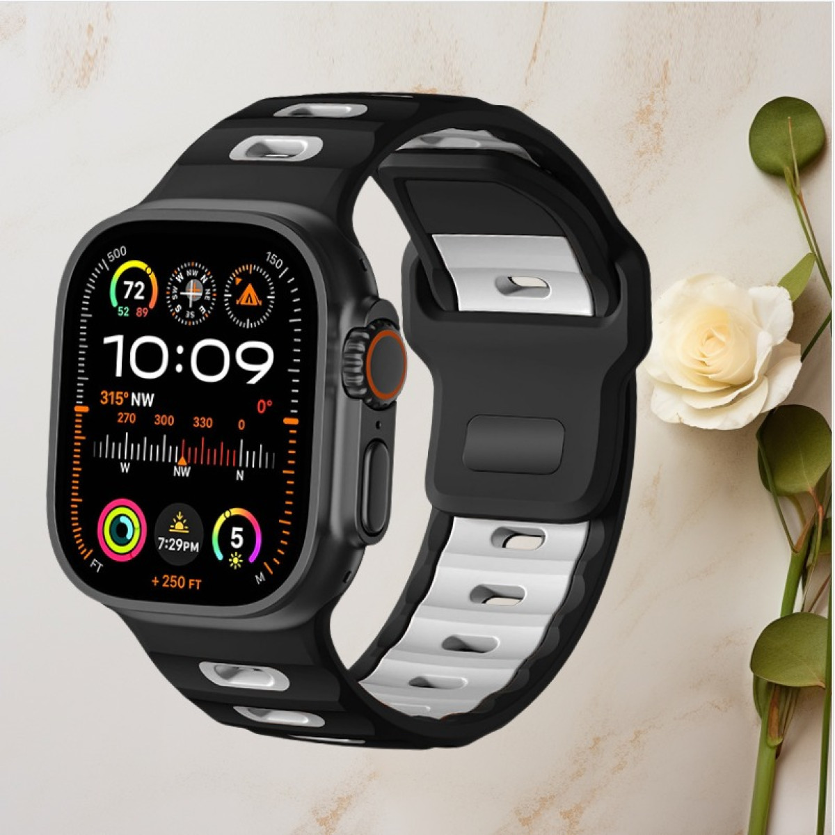 Smartwatch nero con cinturino bianco e nero che mostra l'ora. Fiore bianco e foglie accanto.