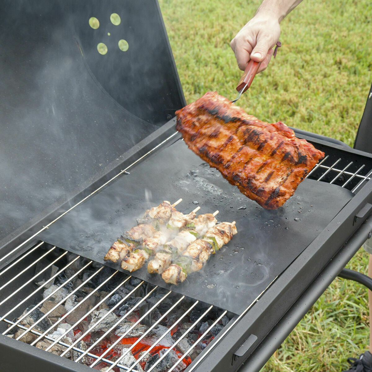 Person grillt Fleisch und Spieße auf einem Grill mit glühenden Kohlen.