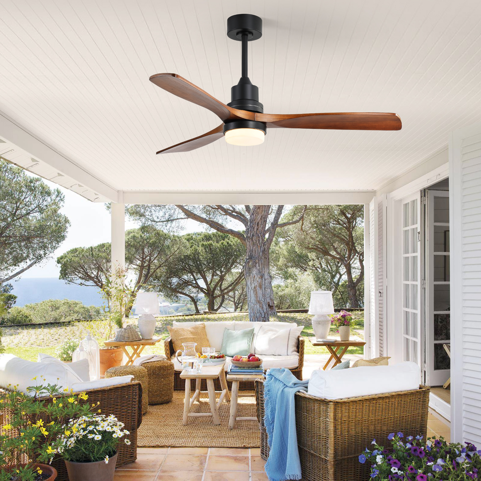 Eine Außenterrasse mit Deckenventilator. Es gibt Korbstühle, ein Sofa und Pflanzen. Das Meer ist im Hintergrund.