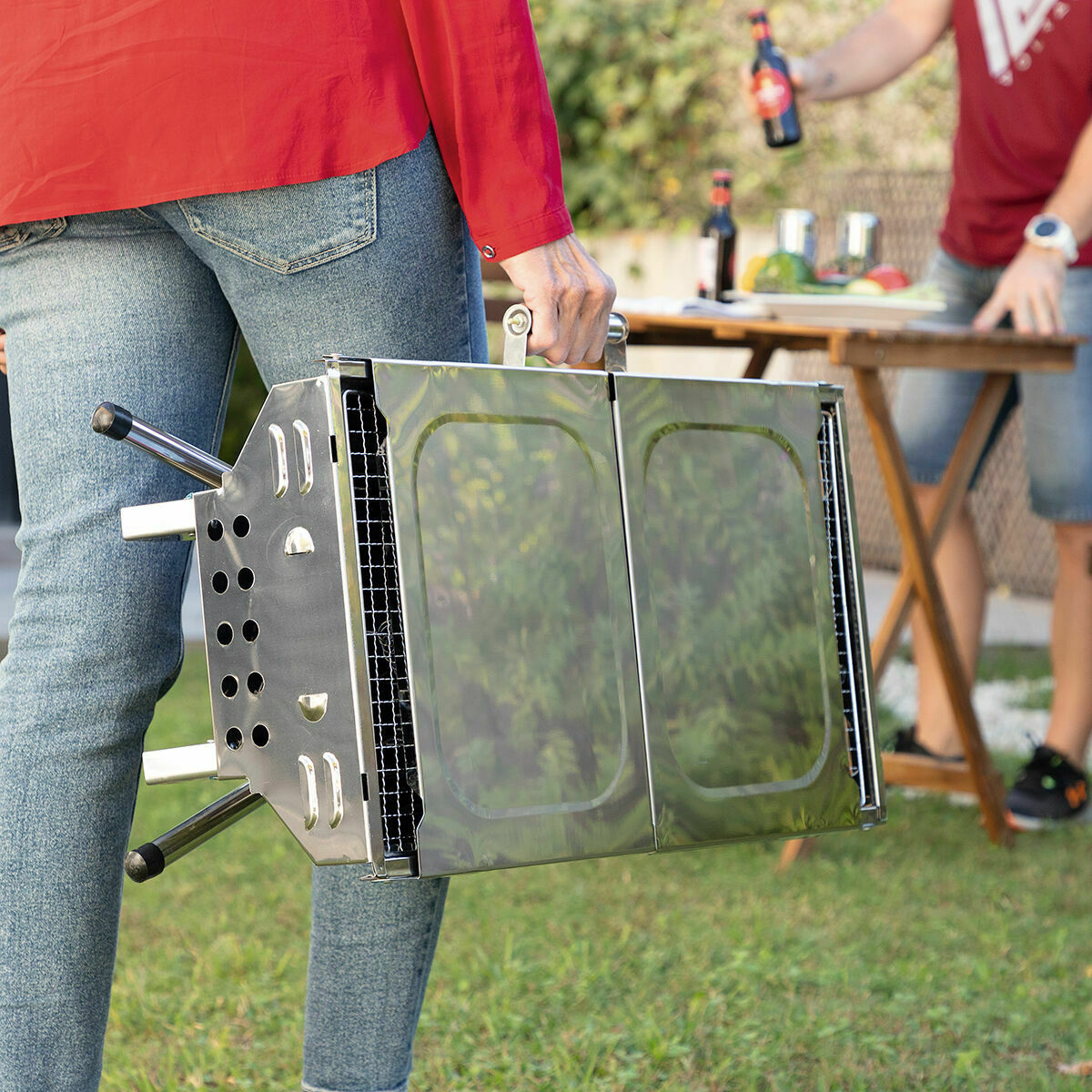 Eine Person trägt einen geschlossenen tragbaren Grill. Eine andere Person ist im Hintergrund in der Nähe eines Tisches.