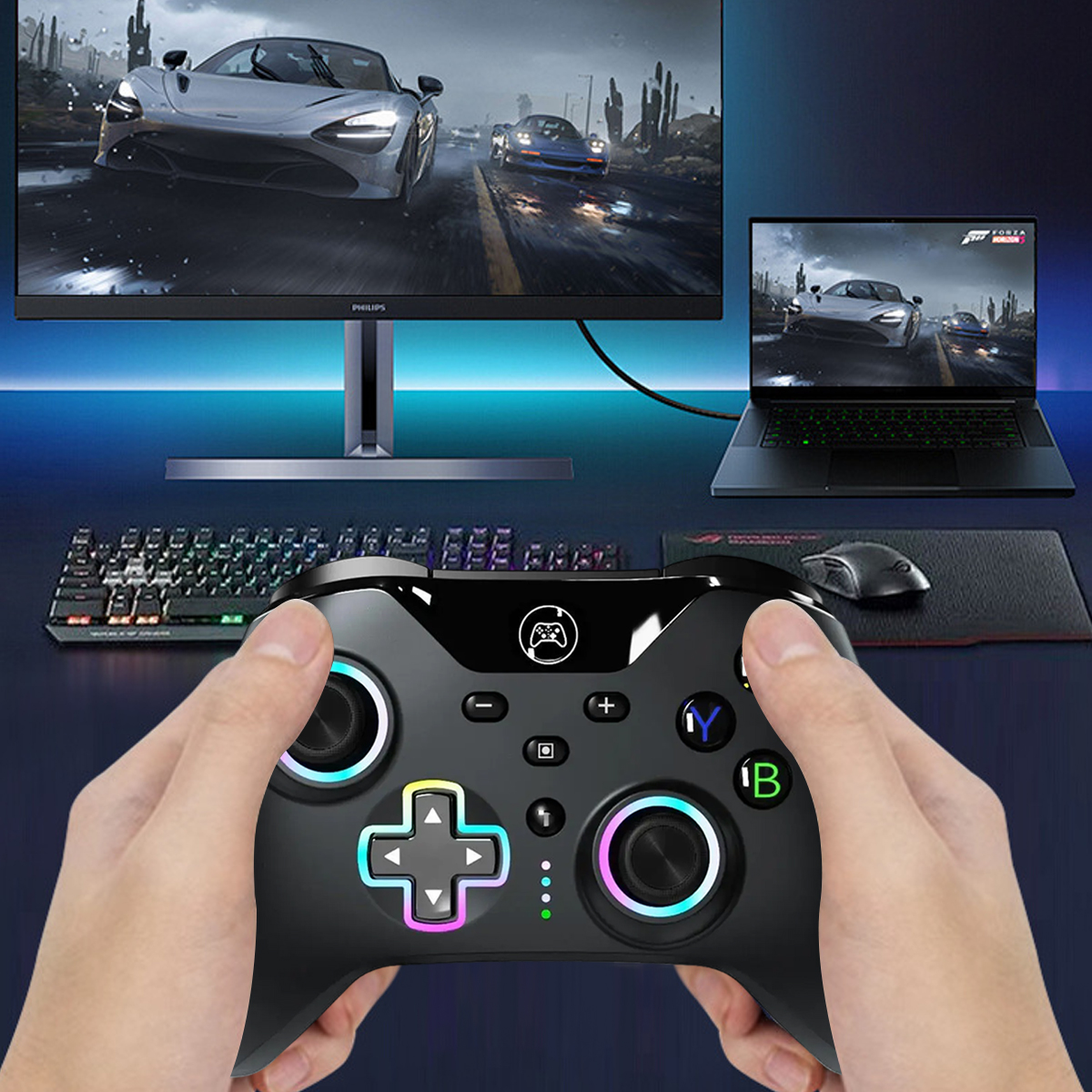 Hände halten einen schwarzen Controller vor einem Computer-Setup. Monitor, Laptop, Tastatur und Maus sind sichtbar.