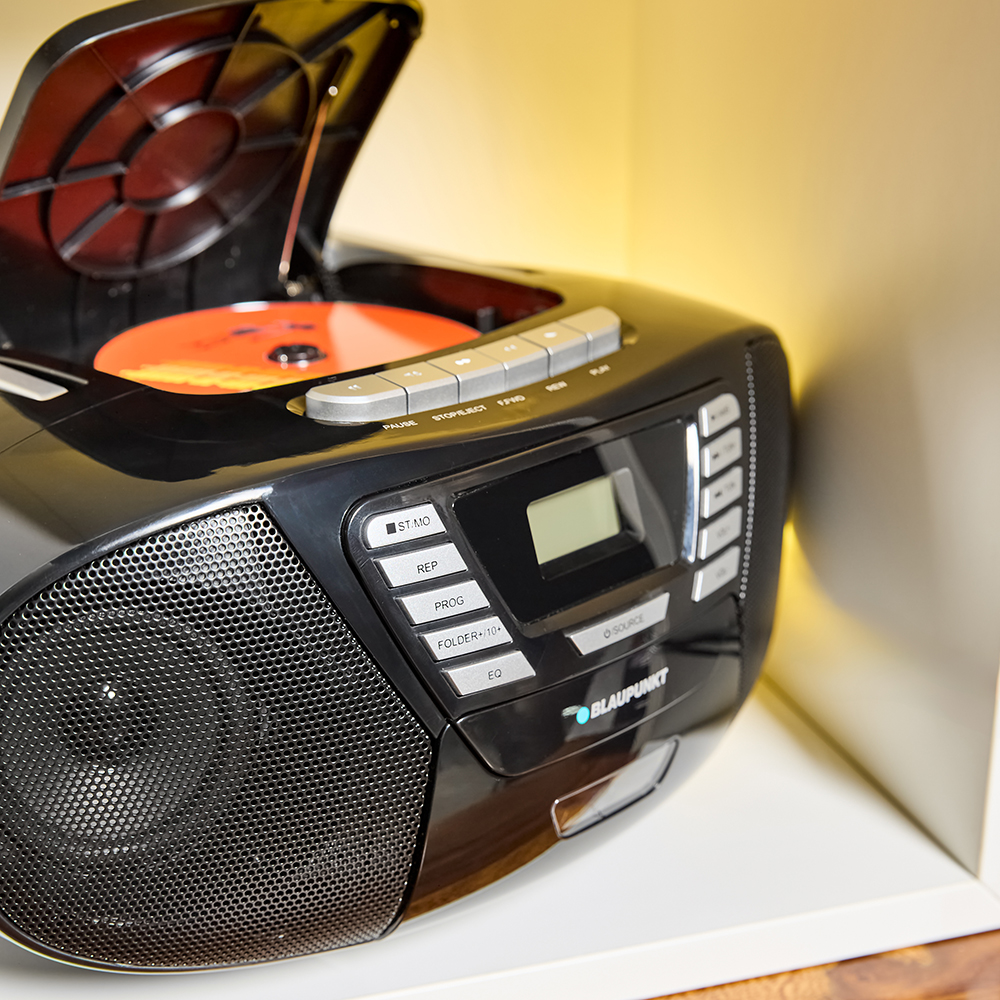 Boombox nero, lettore CD aperto, su una mensola bianca. Ha pulsanti e un display, illuminato da una luce calda.