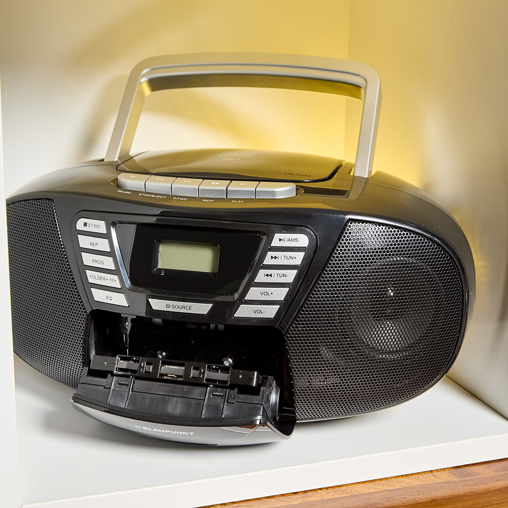 Boombox nero, lettore di cassette aperto, su uno scaffale. Ha display, pulsanti e una maniglia. Illuminato da una luce calda.