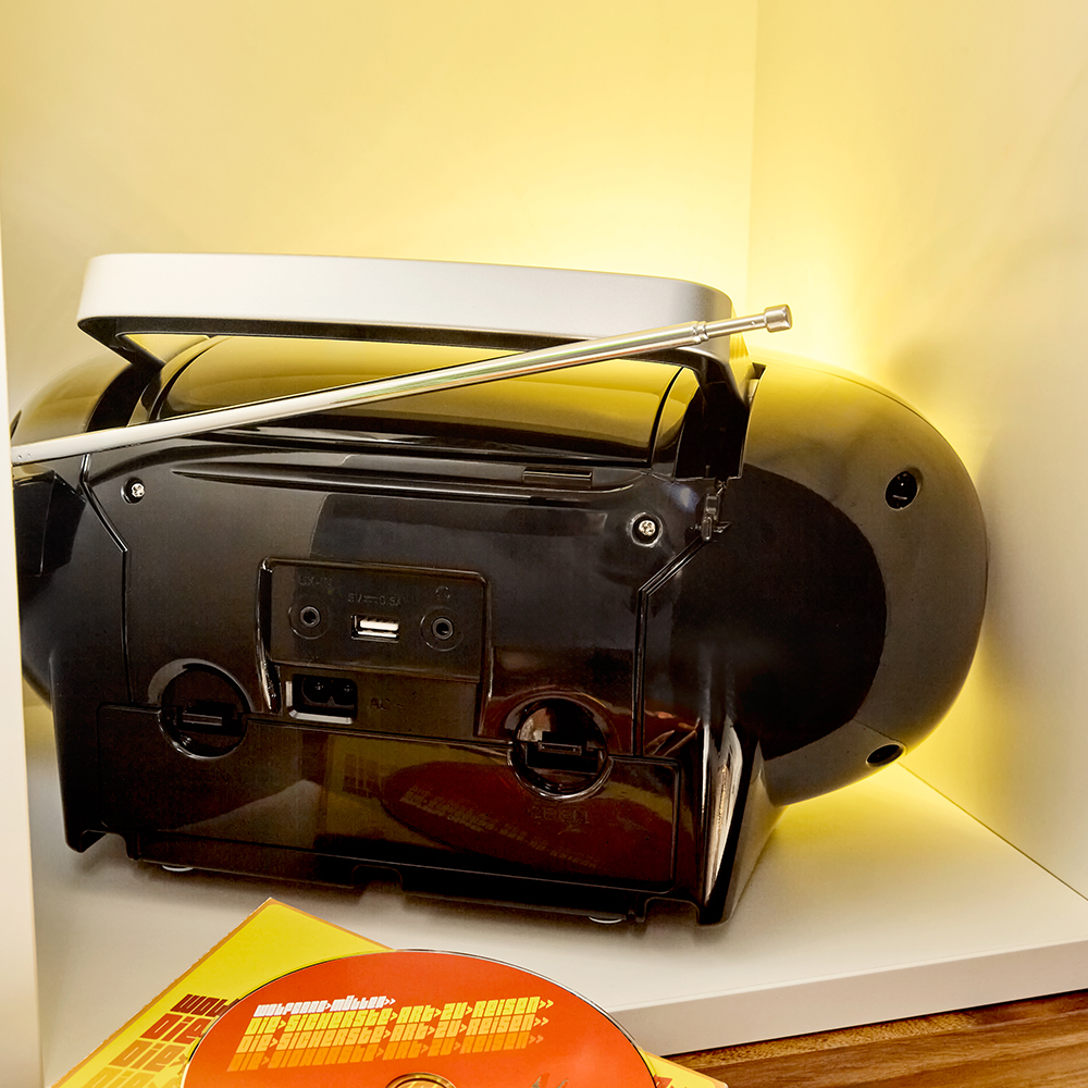 Boombox nero su uno scaffale. Ha un lettore CD, un'antenna e vari pulsanti. Illuminato da una luce calda.