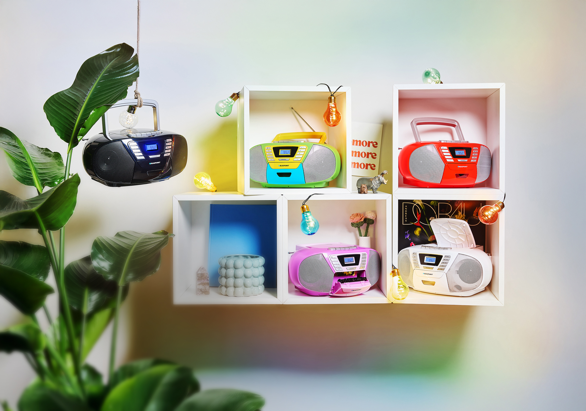 Mehrere Ghettoblaster auf Regalen, jeweils in verschiedenen Farben. Dekoriert mit Lichterketten und Pflanzen.