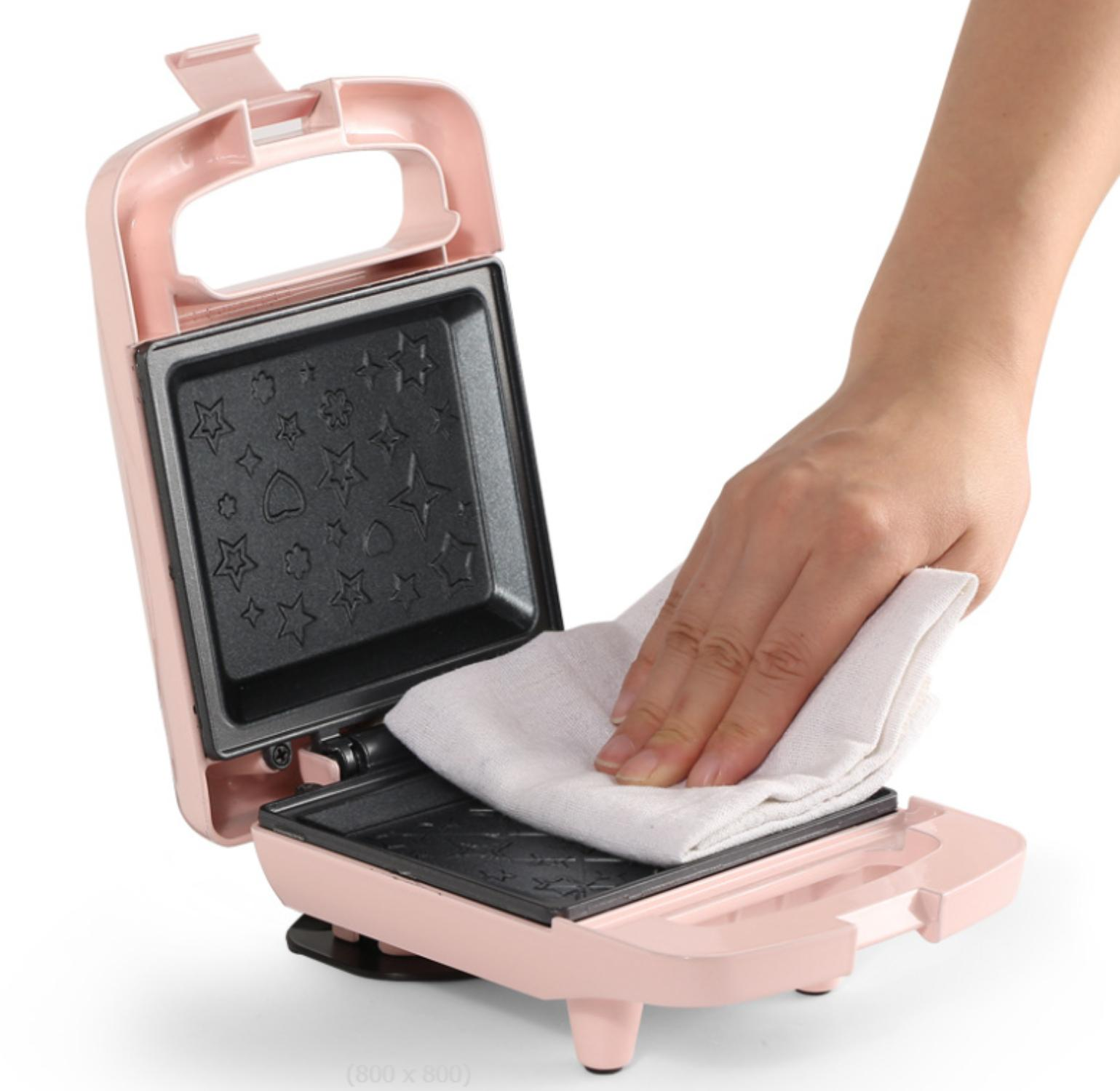 Ein pinker Waffeleisen ist geöffnet. Eine Hand wischt die Innenseite mit einem weißen Tuch ab.