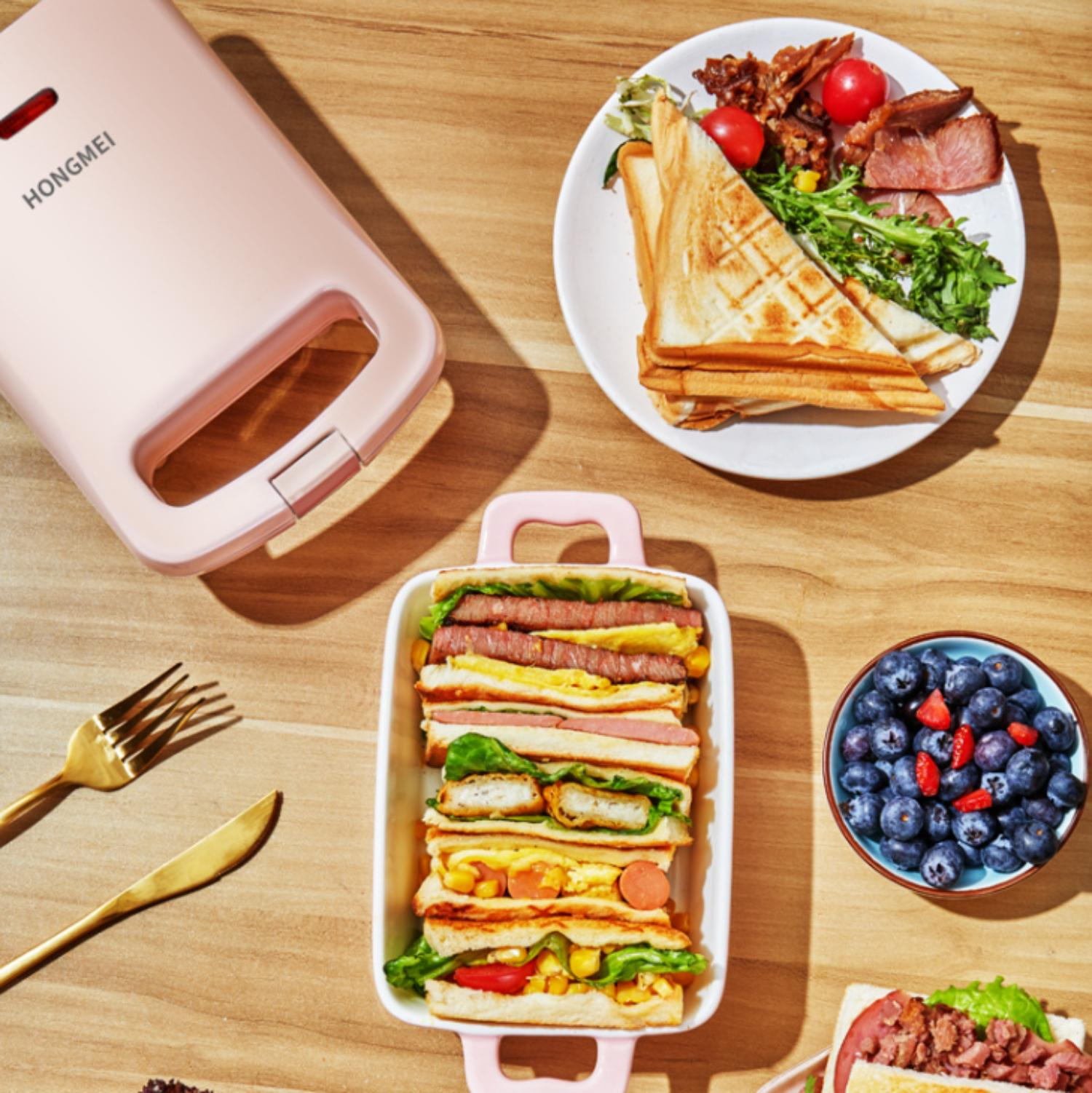 Ein rosa Sandwichmaker neben belegten Sandwiches, einer Obstschale und goldenen Utensilien auf einem Holztisch.