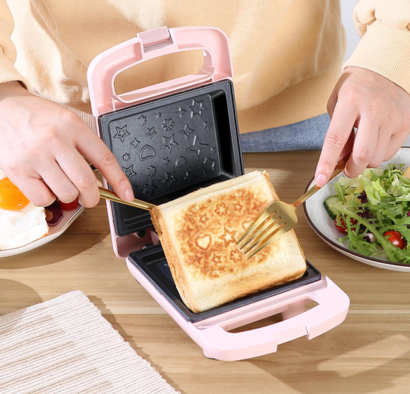 Ein pinkfarbener Waffeleisen, offen, mit einem gerösteten Waffel, die mit goldenen Gabeln angehoben wird.