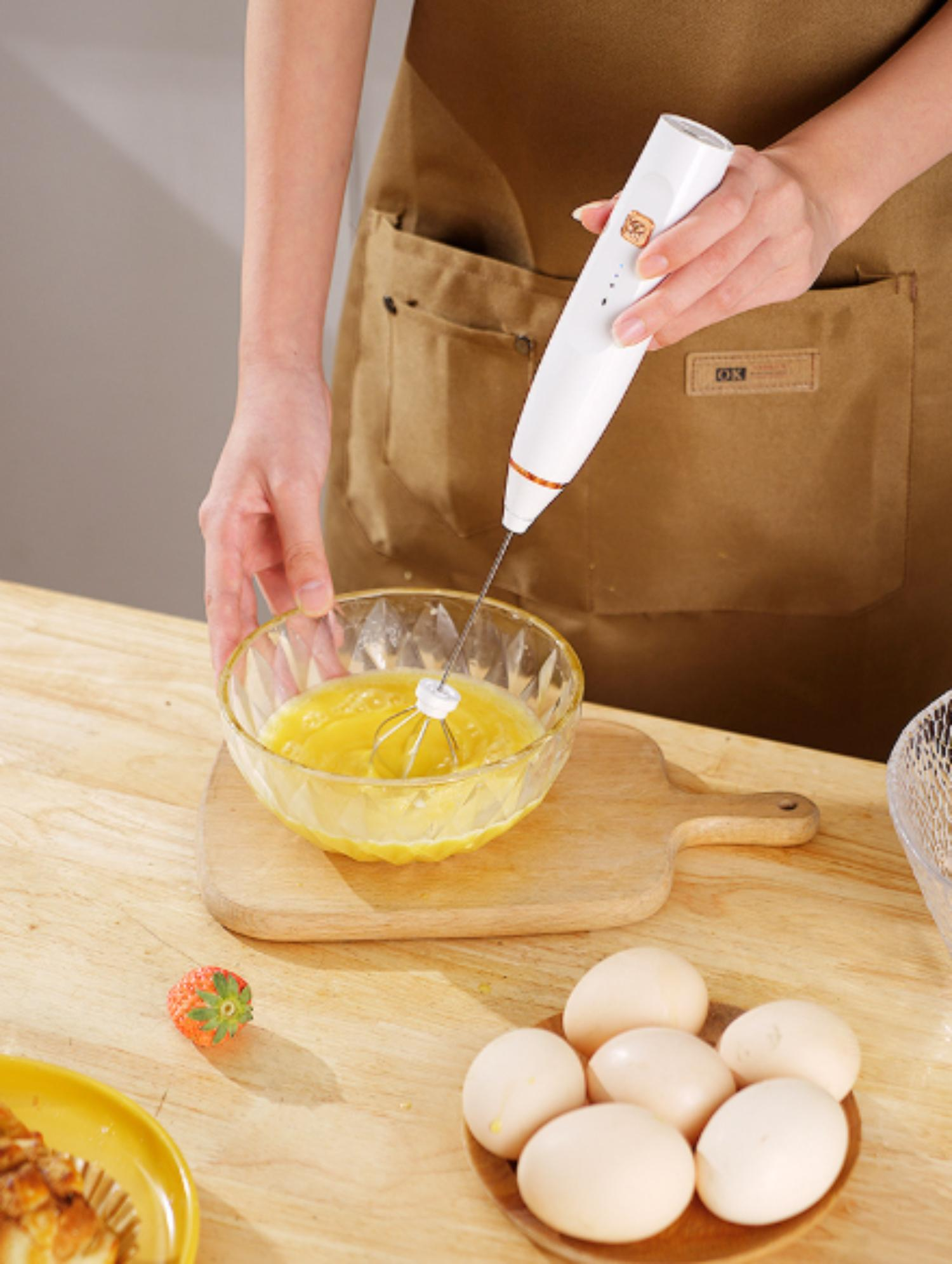 Eine Person verwendet einen weißen und goldenen Handmixer, um Eier in einer Glasschüssel auf einem Holzbrett zu verquirlen.