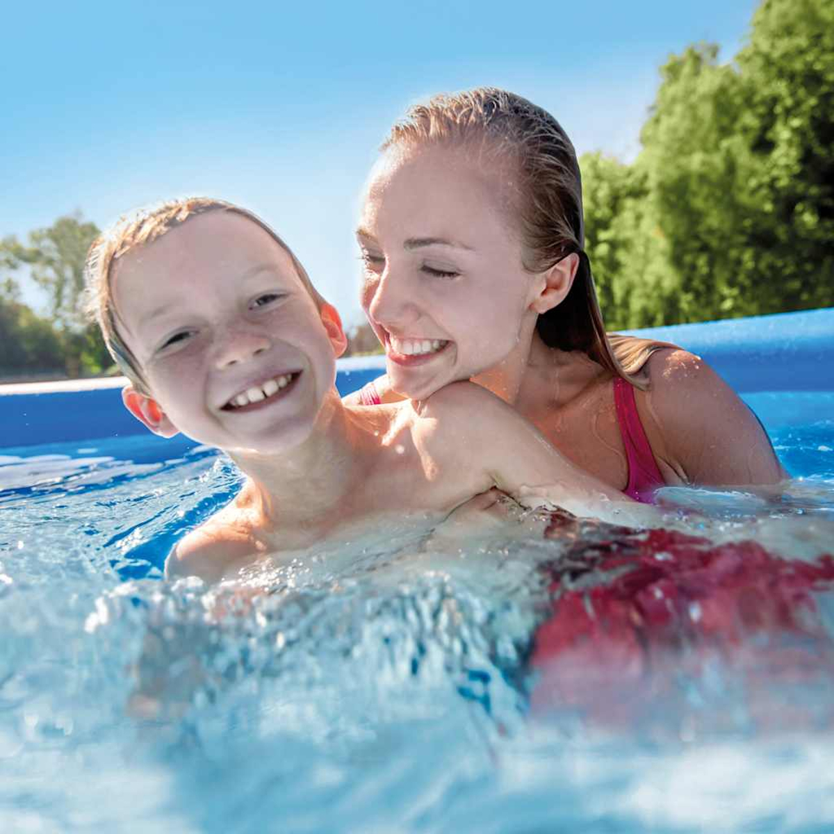 Mujer y niño sonriendo en una piscina azul, salpicaduras de agua, con árboles verdes y cielo azul.