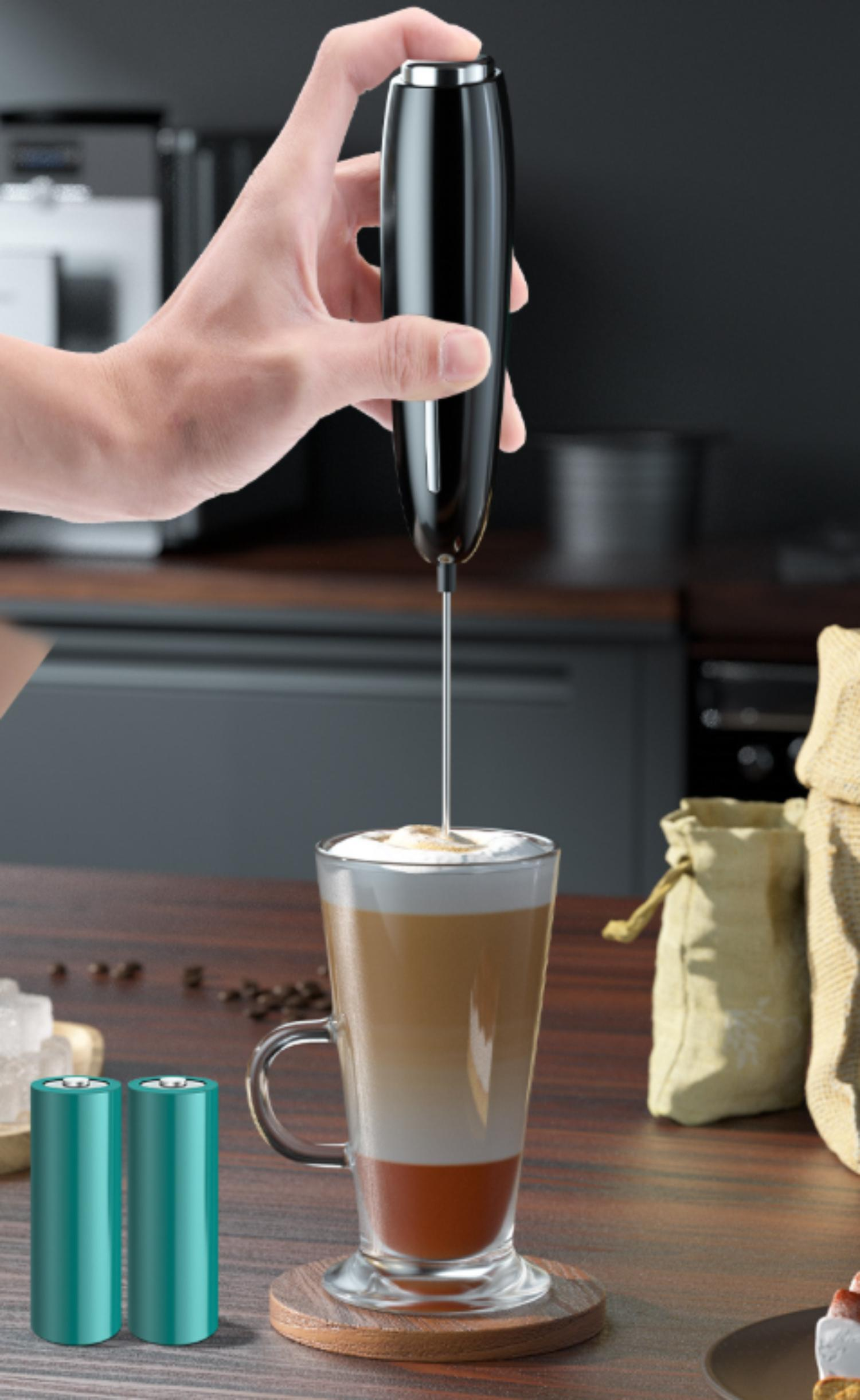Hand hält Milchaufschäumer, schäumt Milch in ein Latte-Glas, zwei grüne Batterien links.