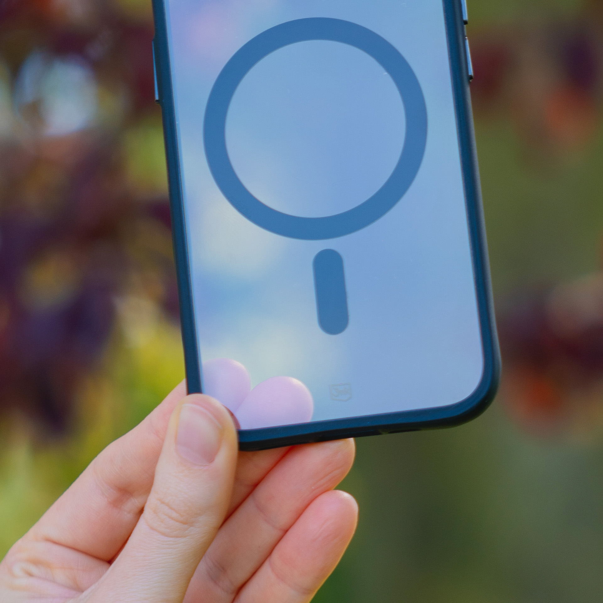 Dłoń trzyma przezroczyste etui na telefon z czarnym kółkiem MagSafe i pionową linią.