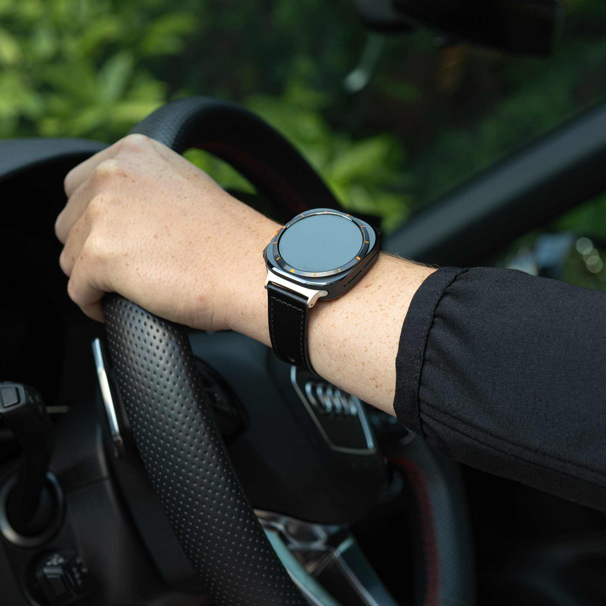 Eine Hand, die eine schwarze Smartwatch trägt, während sie ein Lenkrad in einem Auto hält.