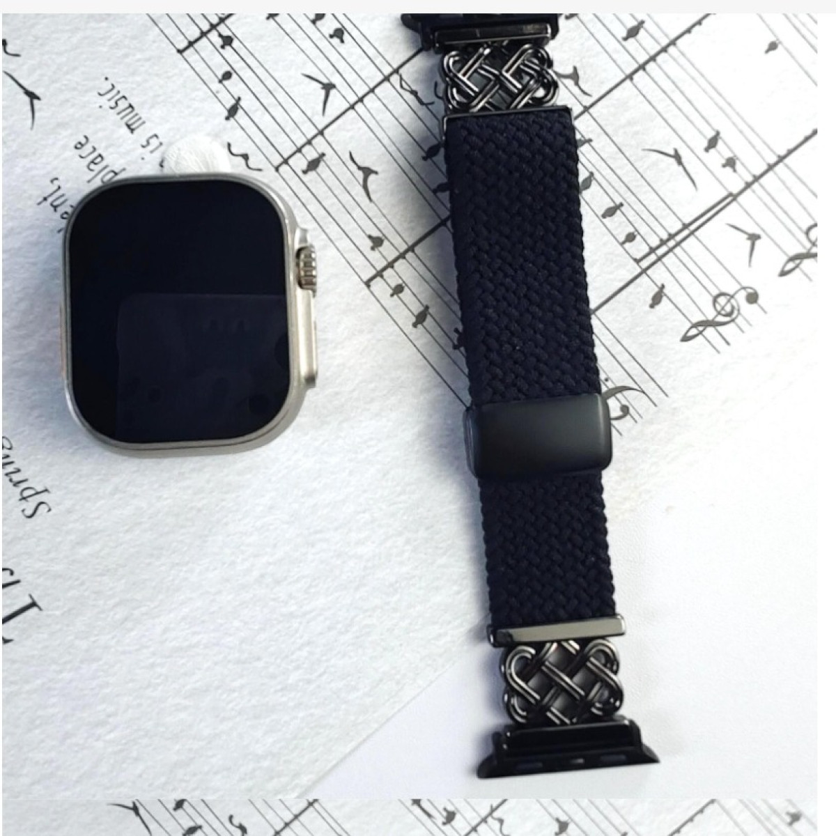 Smartwatch und Armband auf Notenblatt-Hintergrund. Uhr ist silber, Armband dunkelblau mit Silberakzenten.