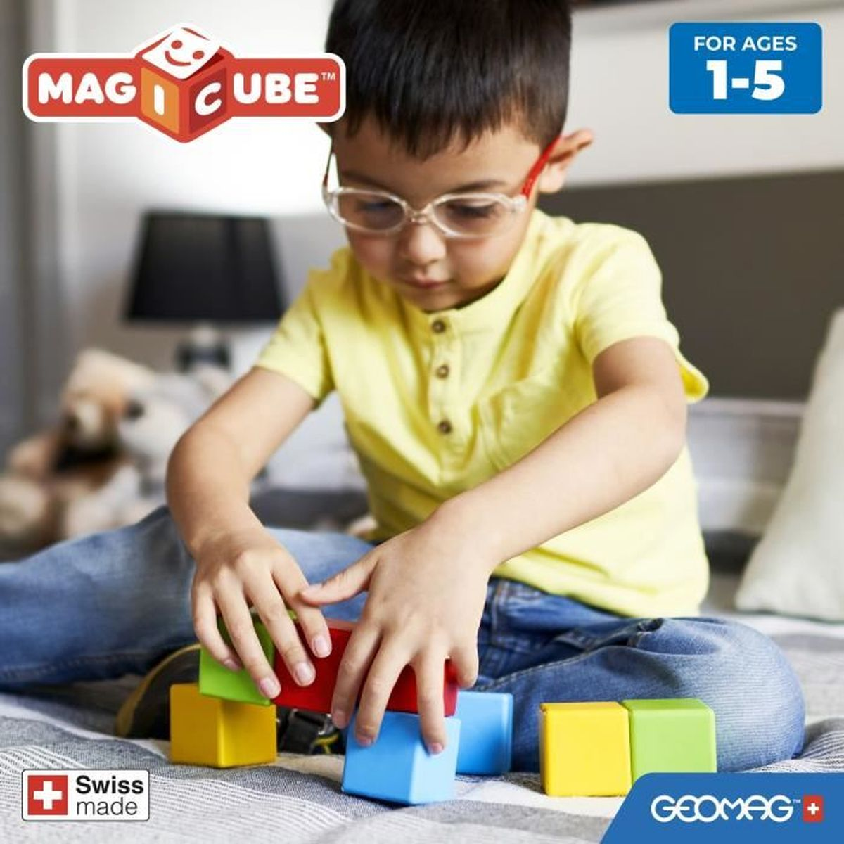 Un bambino che gioca con cubi magnetici. Il bambino indossa occhiali ed è circondato da blocchi. Per età 1-5.