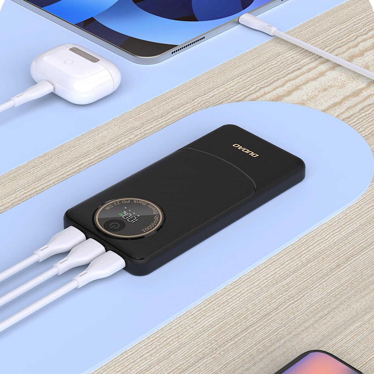 Power bank, AirPods i tablet w trakcie ładowania. Kable łączą urządzenia. Niebieska powierzchnia i drewniany stół.