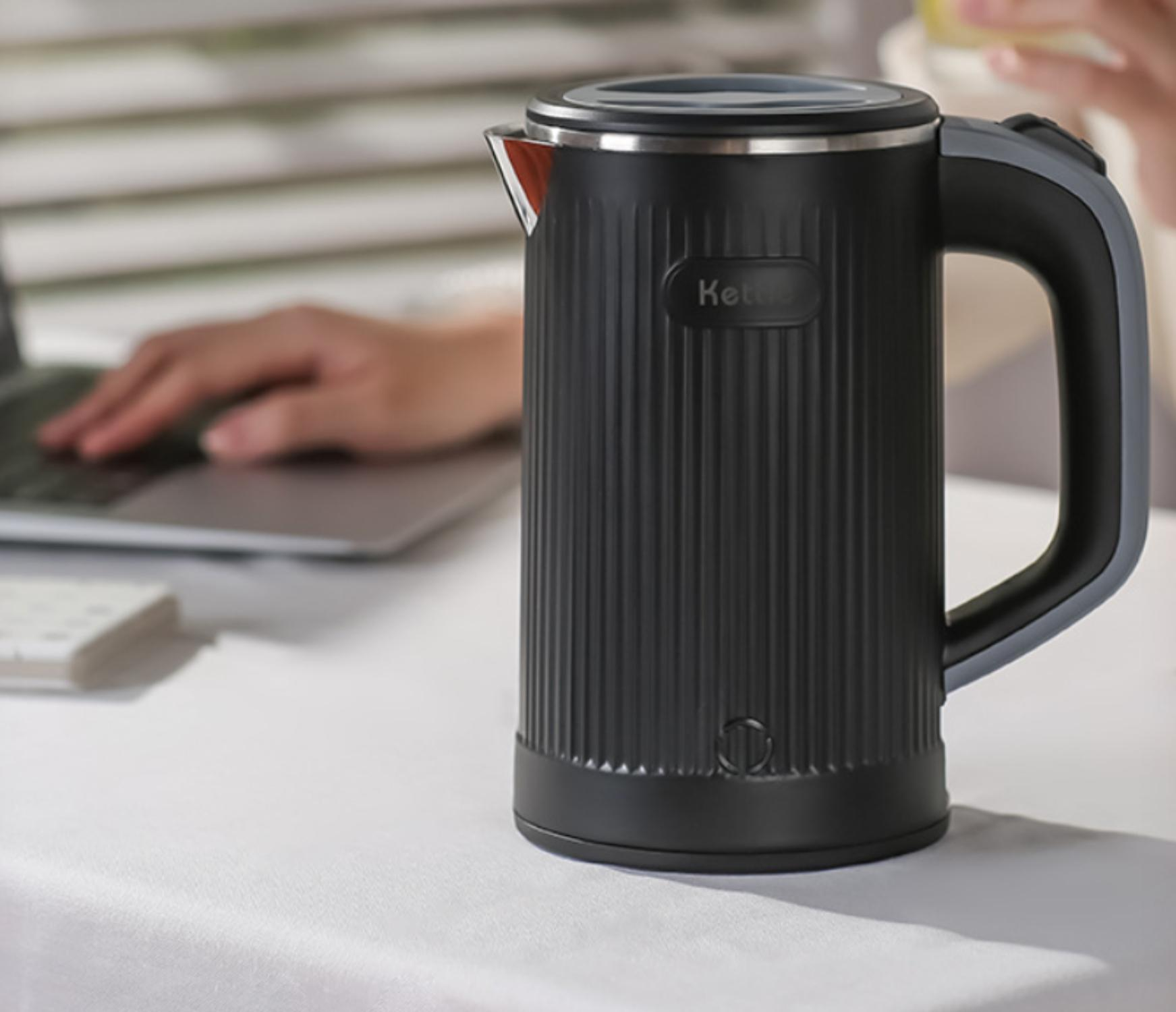 Schwarzer elektrischer Wasserkocher mit geripptem Design, Griff und weißer Oberfläche, platziert in der Nähe von Laptop und Tastatur.