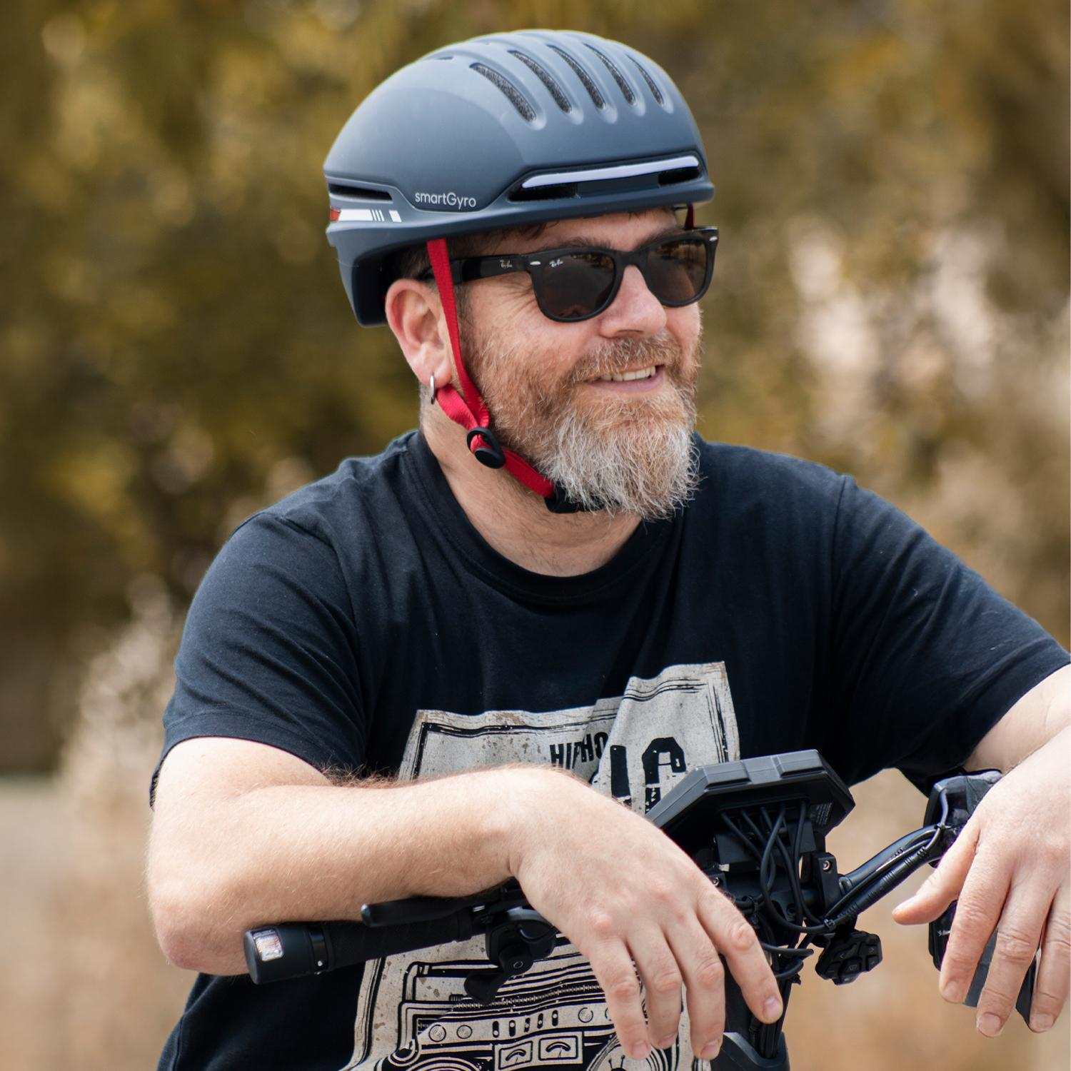 Hombre con casco y gafas de sol. Sonríe, sujetando el manillar, con árboles detrás.