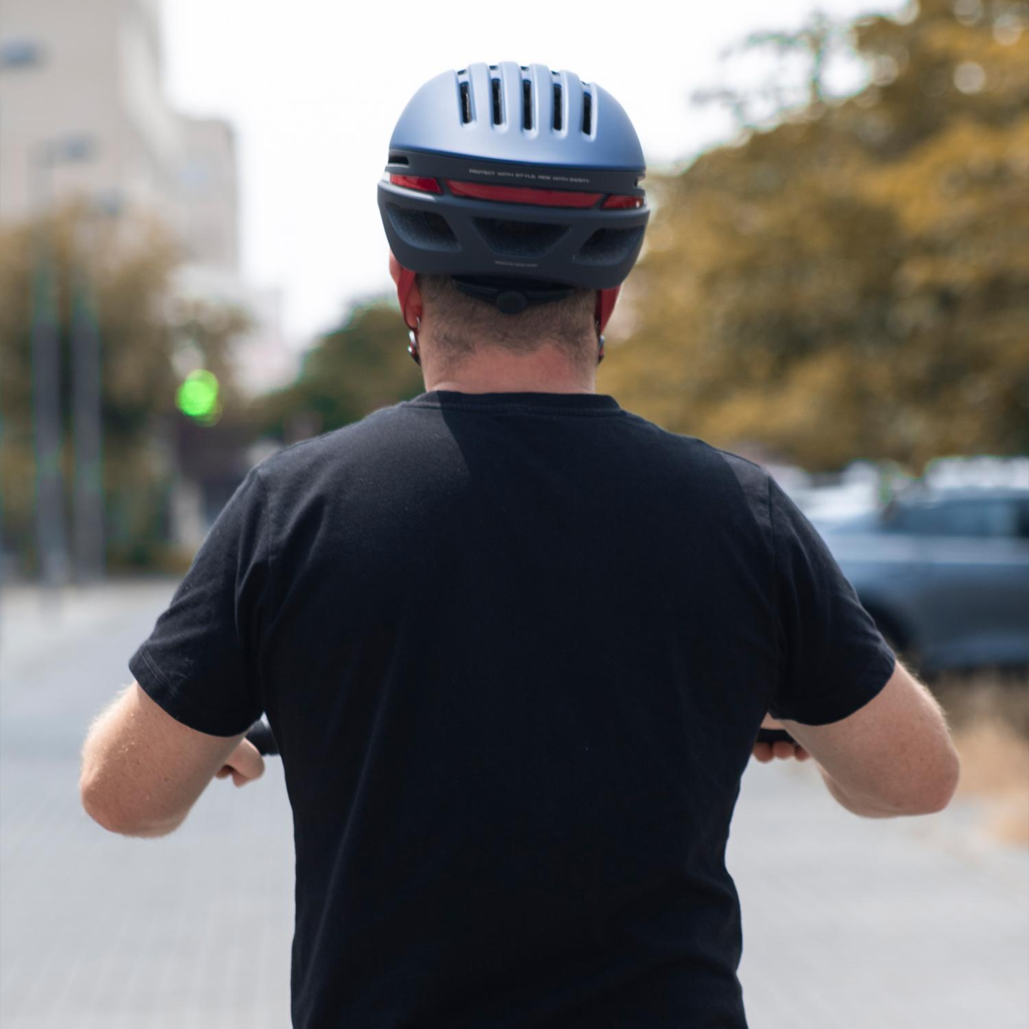 Vista trasera de un hombre con camisa negra y casco azul, montando un scooter en la ciudad.