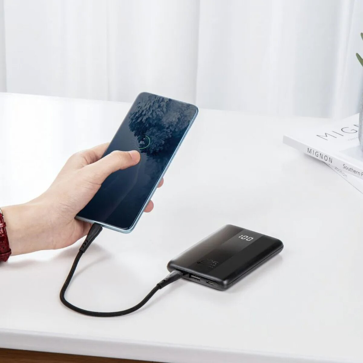 Eine Hand, die ein Telefon hält, das über ein schwarzes Kabel mit einer Powerbank verbunden ist. Ein Schreibtisch mit einem Buch.
