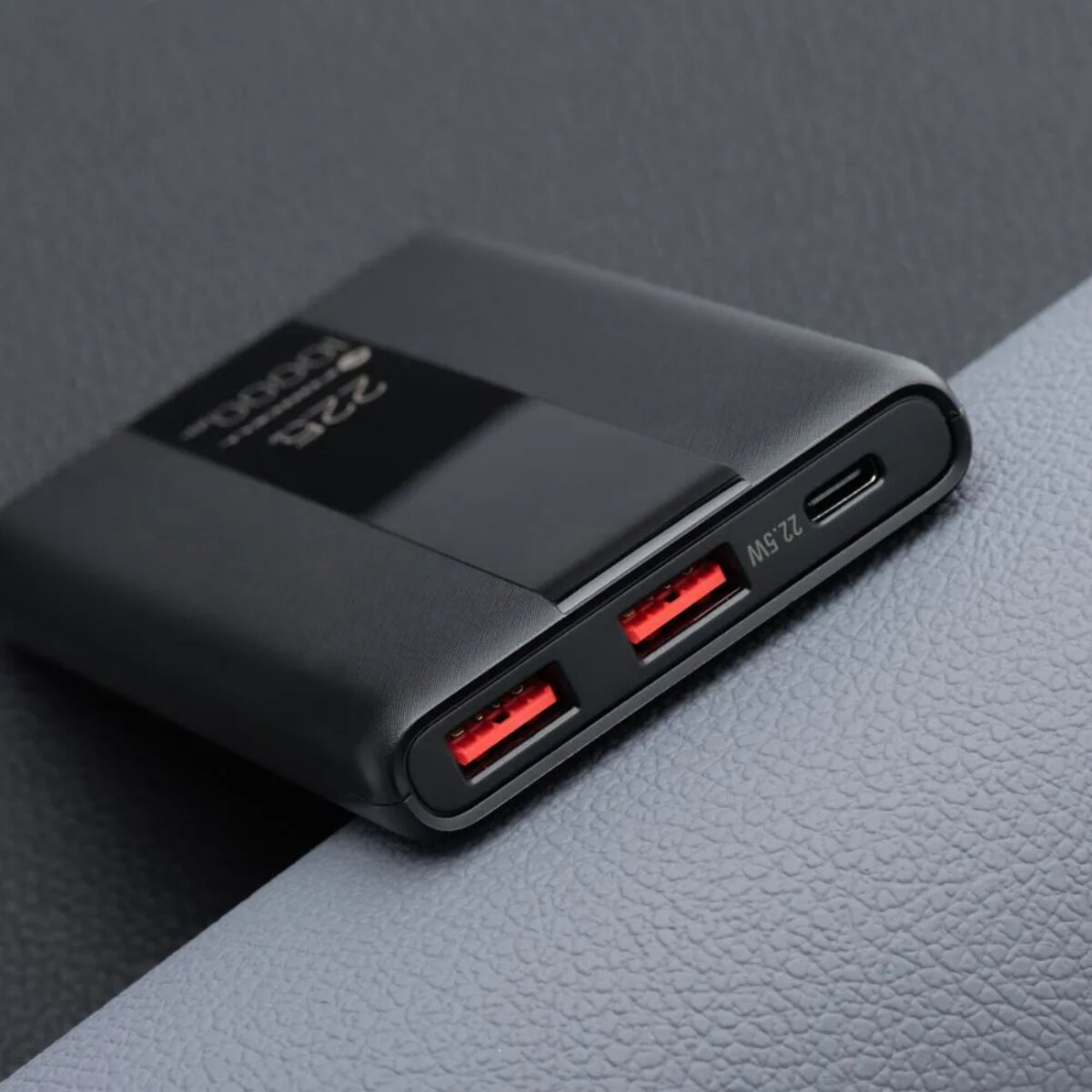Schwarze Powerbank mit zwei roten USB-Anschlüssen und einer Digitalanzeige. Grauer strukturierter Hintergrund.