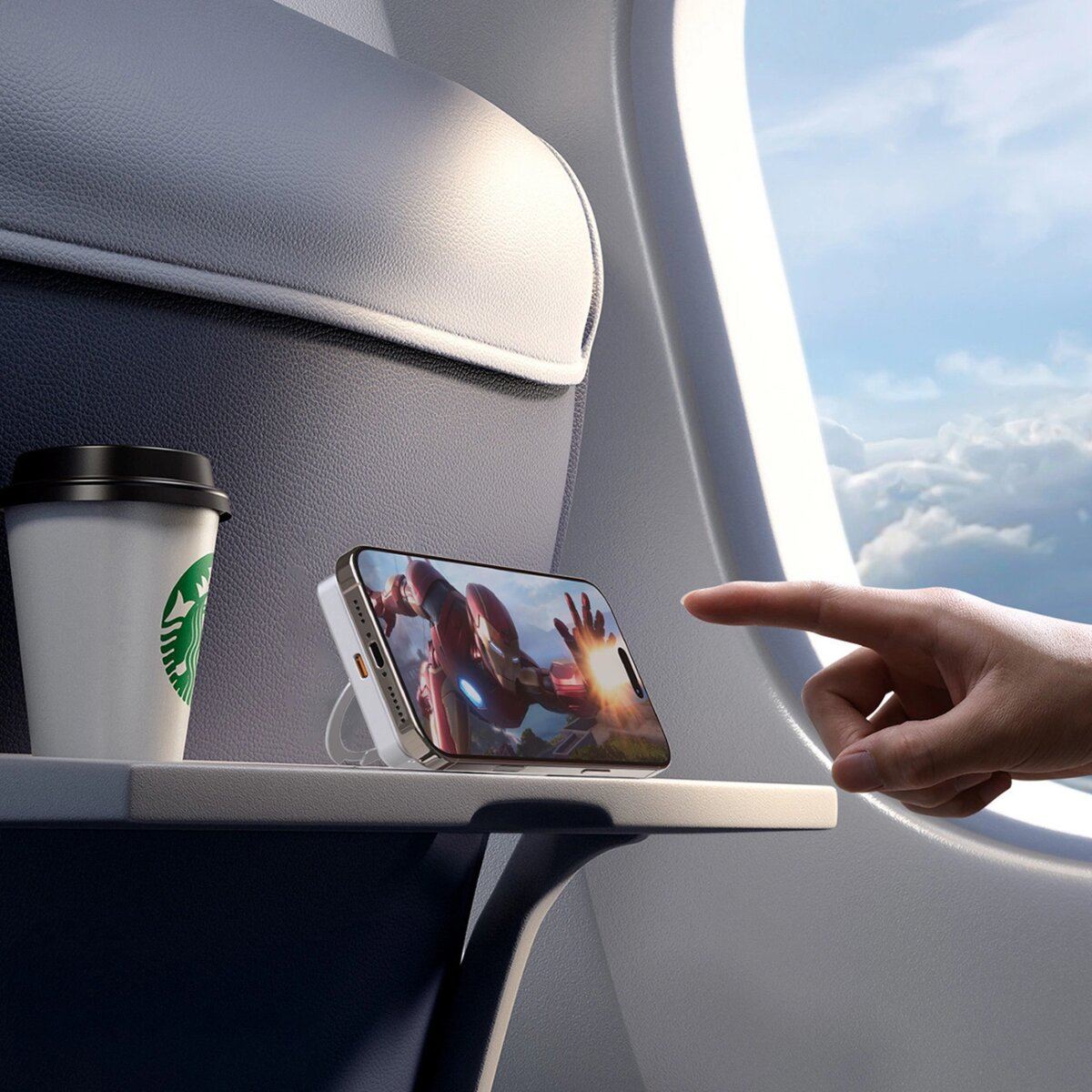 Ręka wskazująca na telefon ładujący się na tacy. Obok kubek Starbucks. Widoczne okno samolotu.