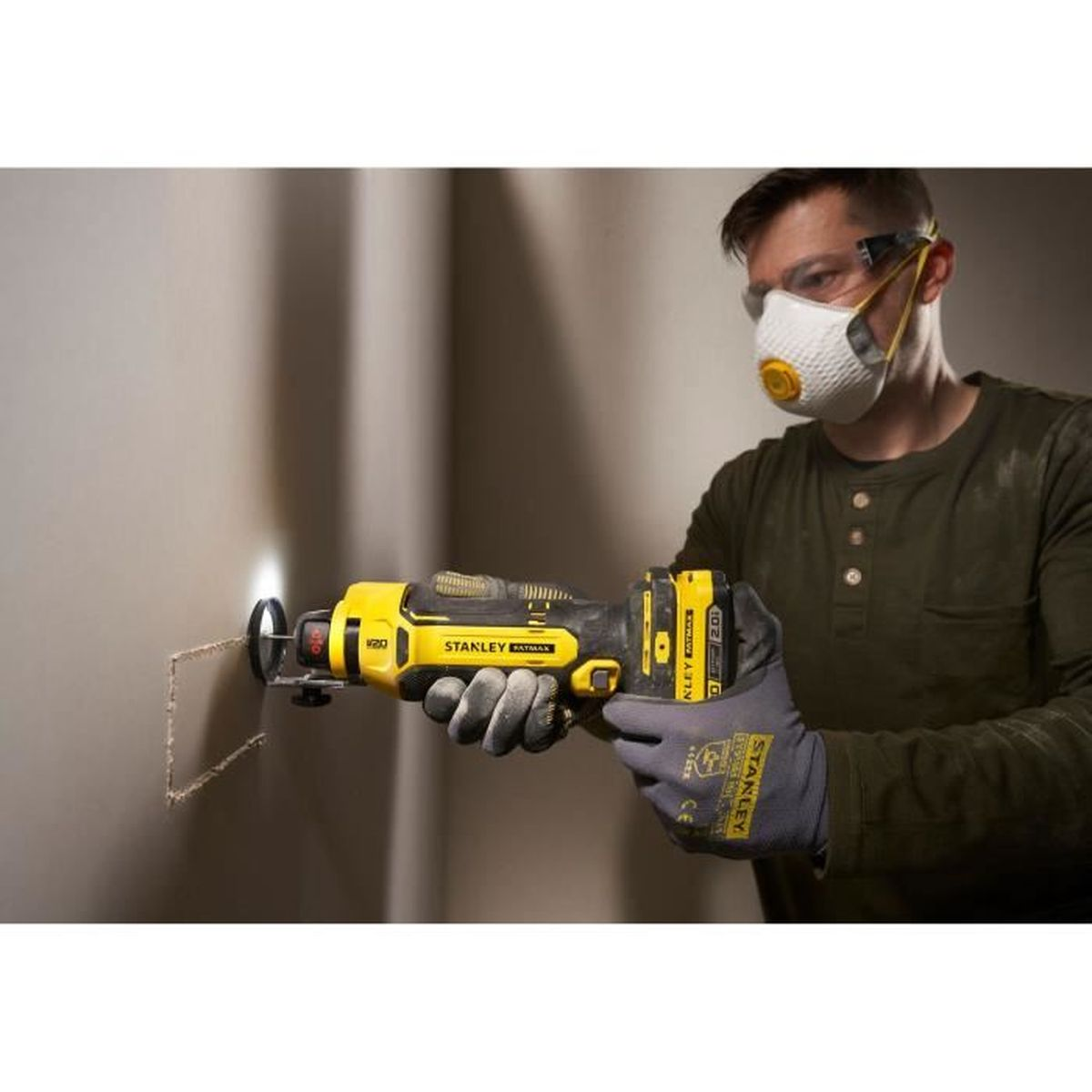 Ein Mann mit Maske und Schutzbrille benutzt ein gelbes Werkzeug, um in eine Wand zu schneiden. Werkzeug ist Stanley Fatmax.