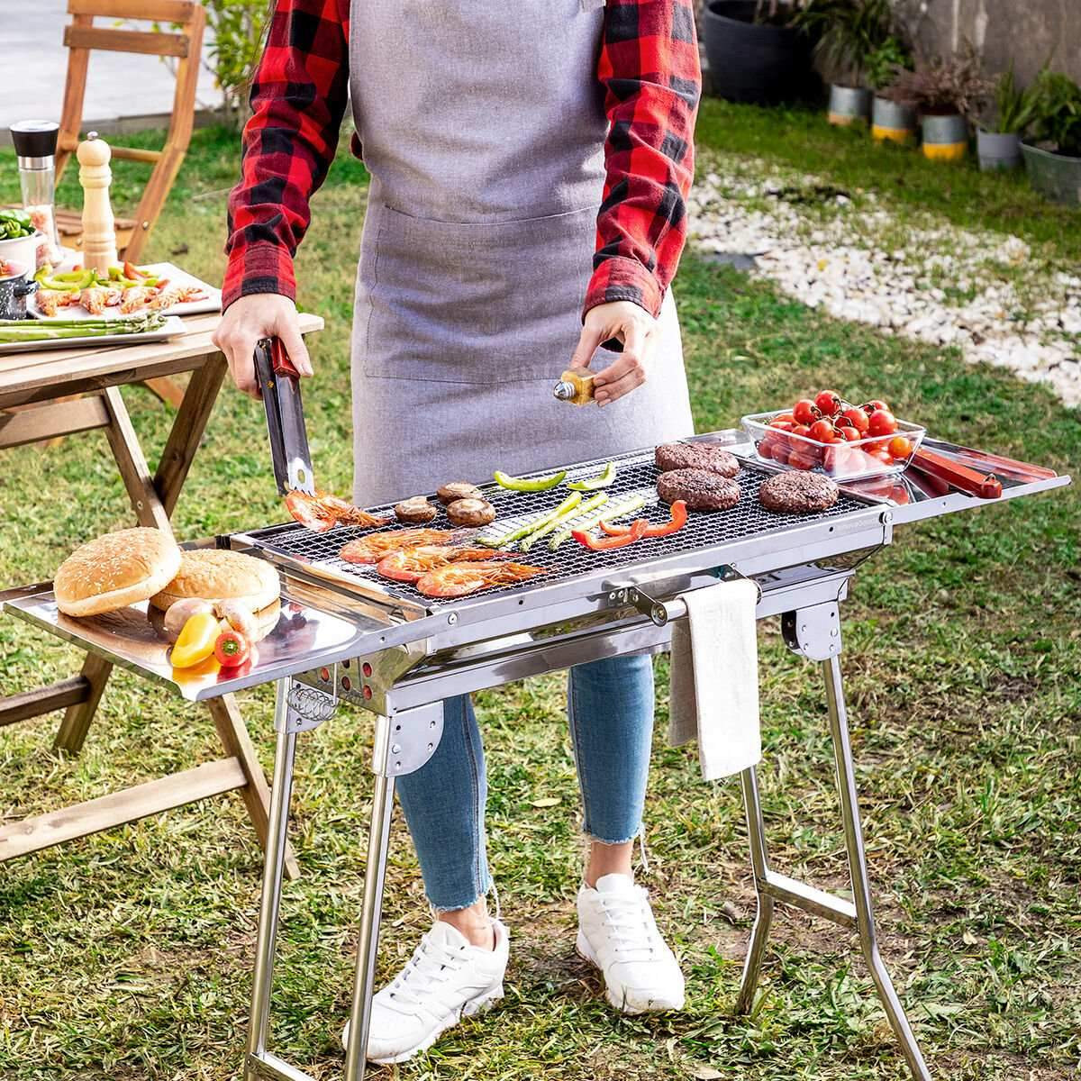 Person, die Essen auf einem tragbaren silbernen Grill mit Beistelltischen in einer Gartenumgebung grillt.