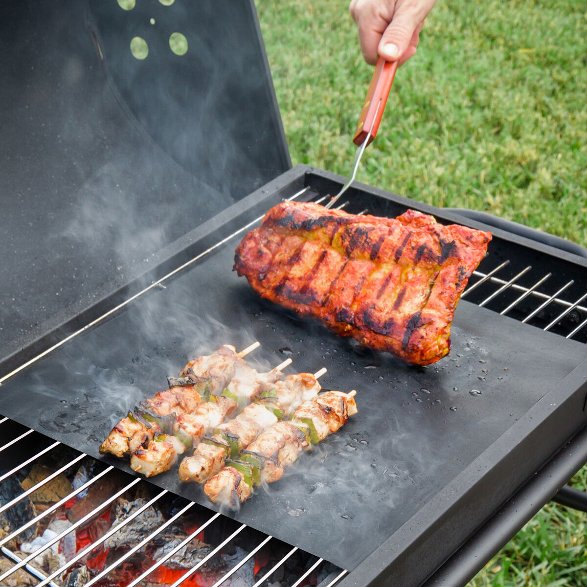 Carne y brochetas a la parrilla sobre una alfombrilla negra en una parrilla. Sale humo.