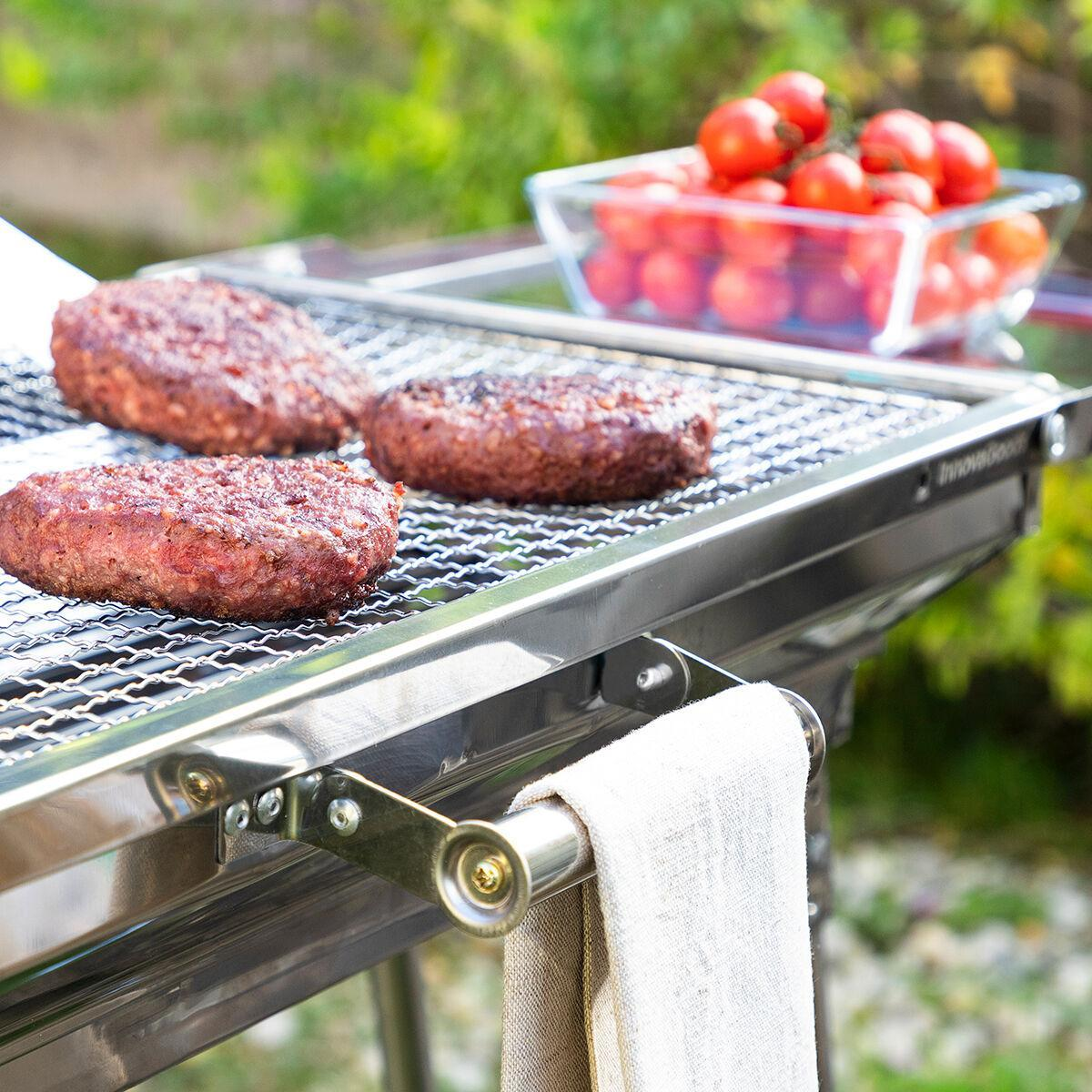 Drei Burger auf einem Grill. Eine Schüssel mit Tomaten im Hintergrund. Darunter hängt ein Handtuch.