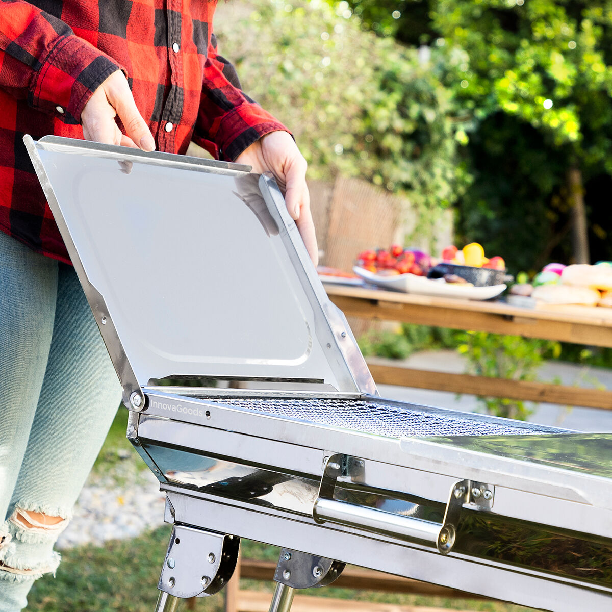 Person öffnet Deckel eines silbernen Grills. Rot-schwarzes kariertes Hemd, Jeans. Draußen.