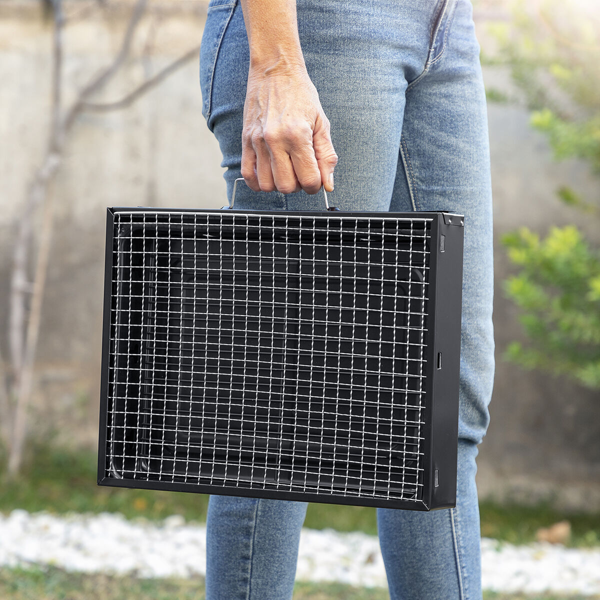 Eine Frau hält einen schwarzen Grill, trägt Jeans, draußen.