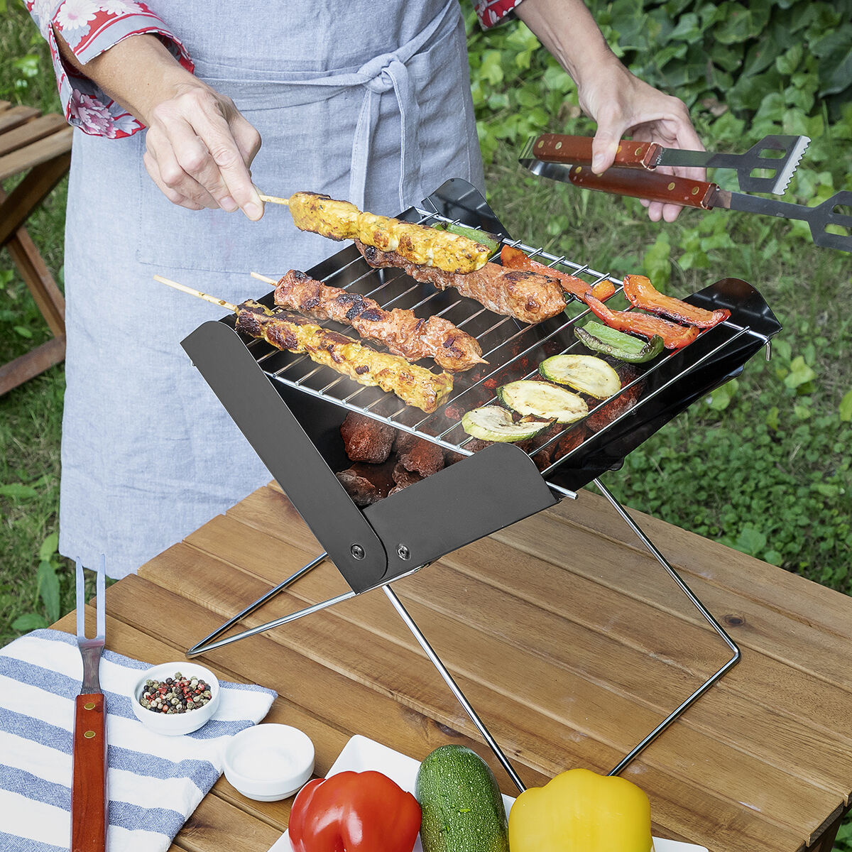 Persona asando brochetas en una parrilla portátil con pinzas, sobre una mesa de madera.