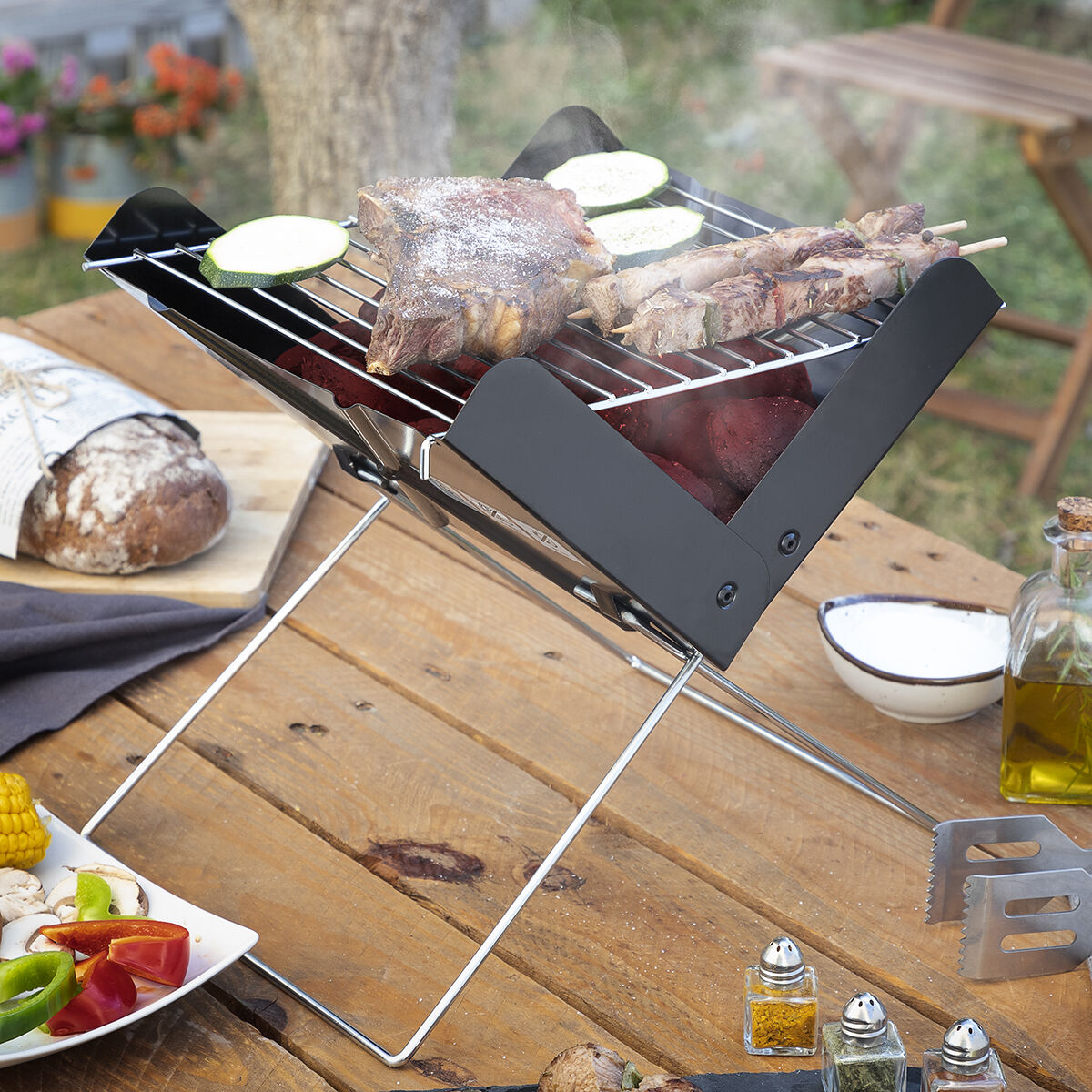 Una parrilla portátil sobre una mesa de madera con carne, verduras y condimentos en un entorno al aire libre.
