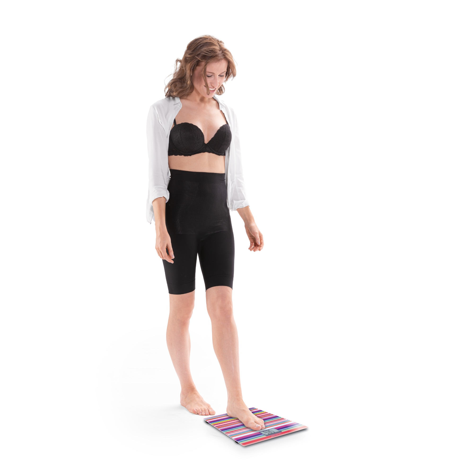Kobieta stoi na wadze w czarnej bieliźnie i białej koszuli. Uśmiecha się.