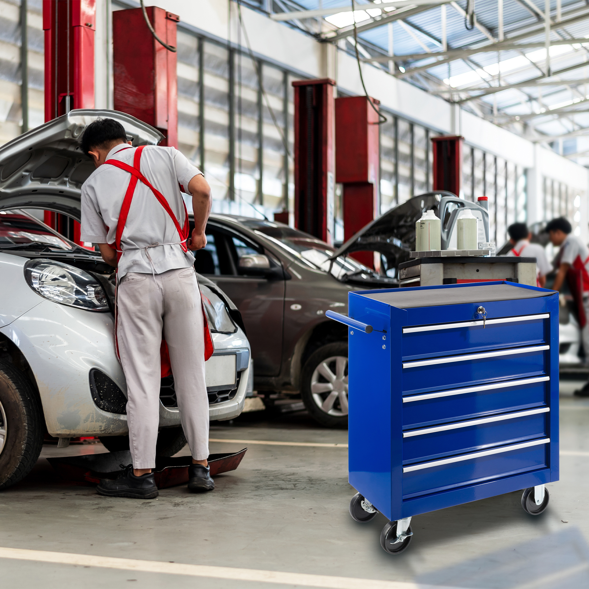 Ein Mechaniker arbeitet in einer Werkstatt an einem Auto, im Vordergrund eine blaue Werkzeugkiste auf Rädern.