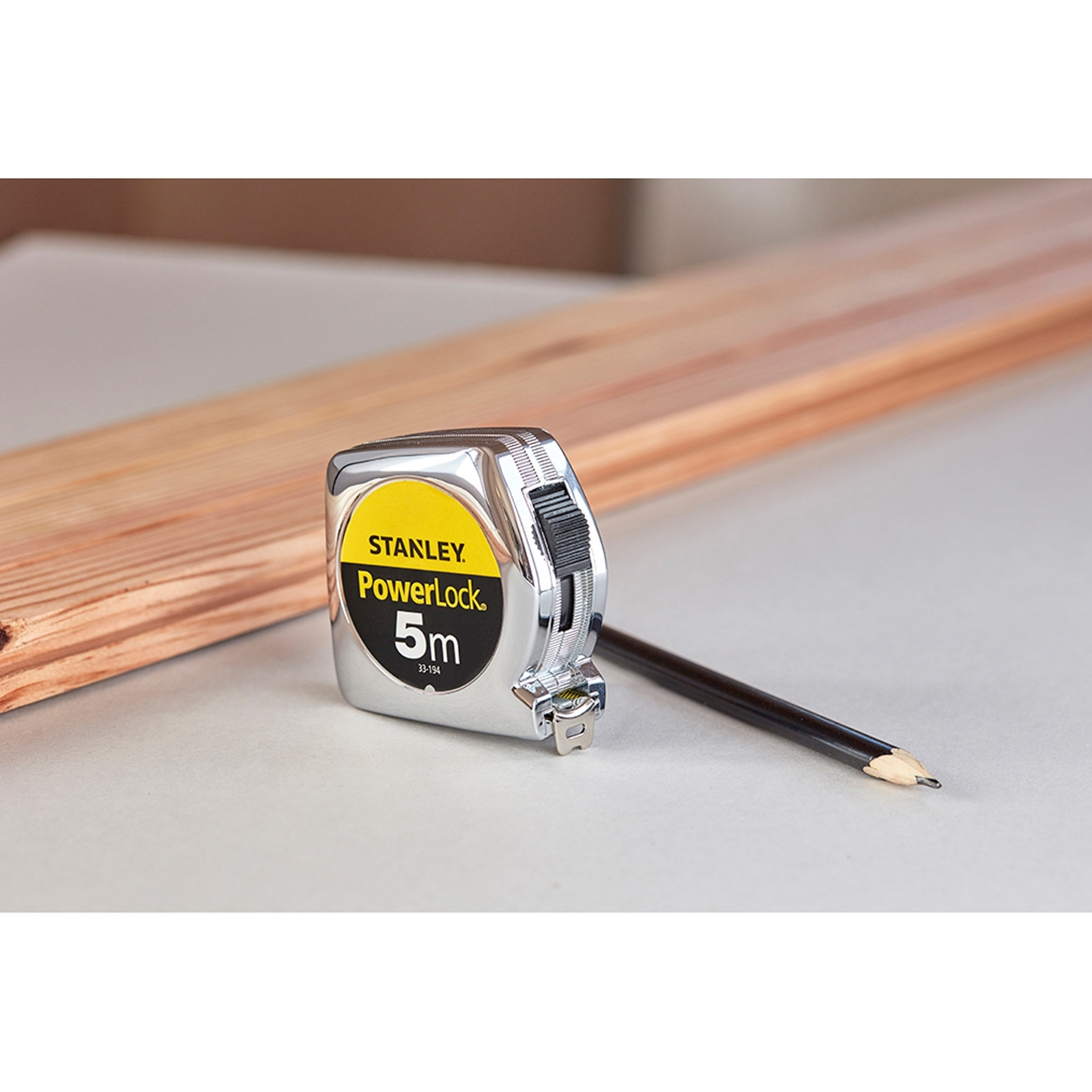 Miarka i ołówek leżą na drewnianej desce. Miarka jest żółta i chromowana.